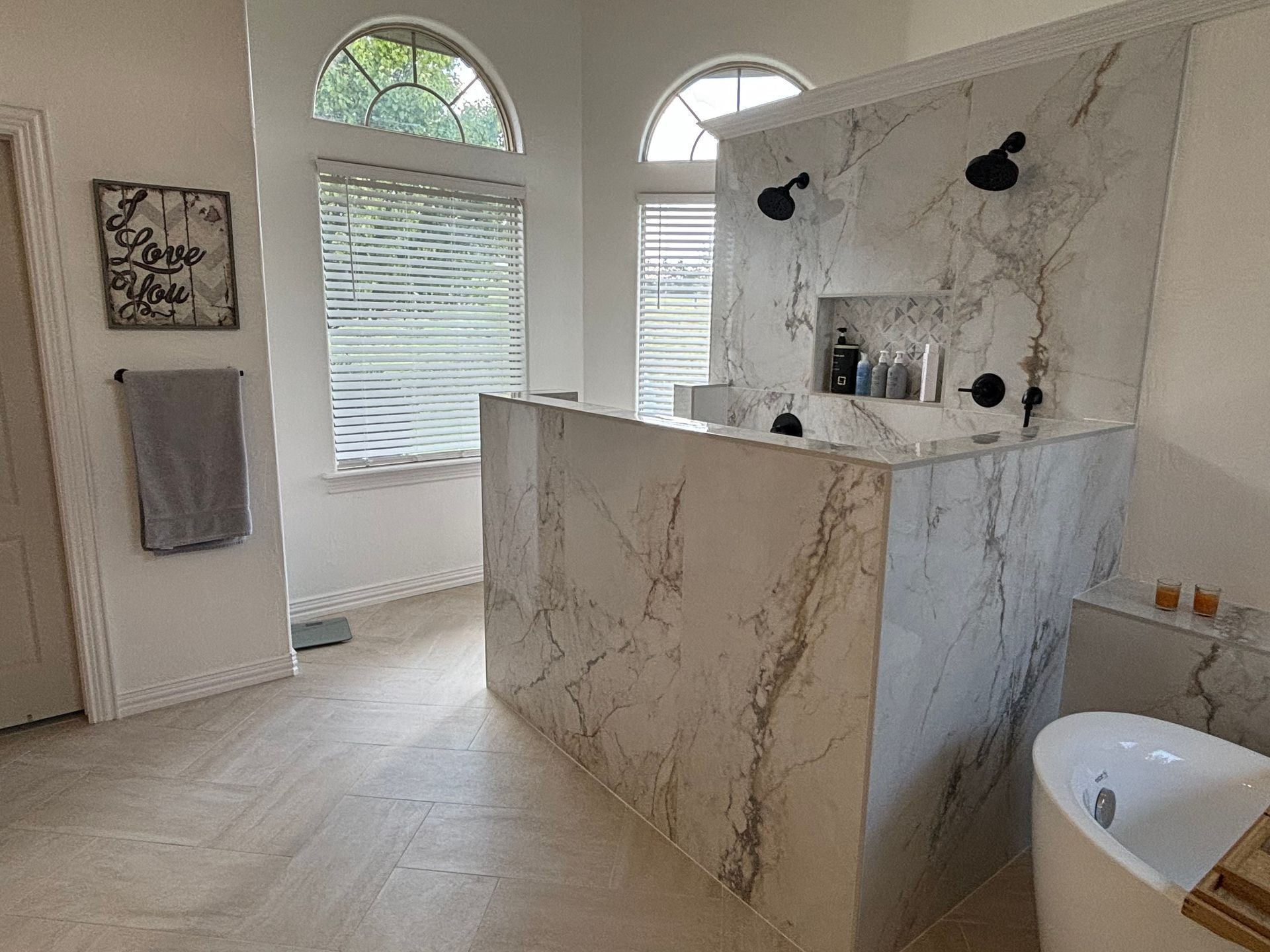 Luxurious bathroom with marble-tiled shower, white walls, arched windows with blinds, and freestanding tub.