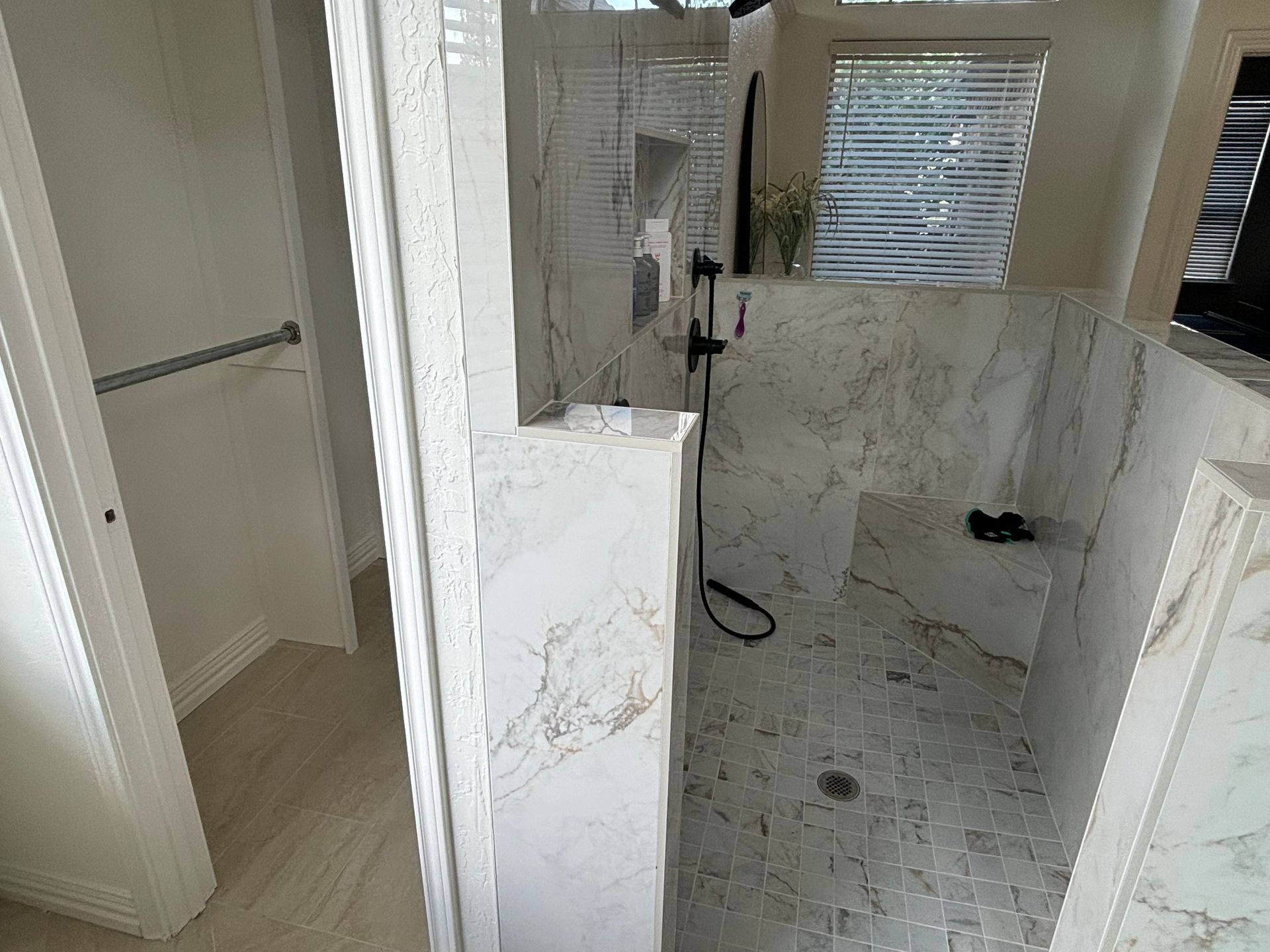 Marble-tiled shower with a bench, black fixtures, and a glass partition. Adjacent is a closet area.