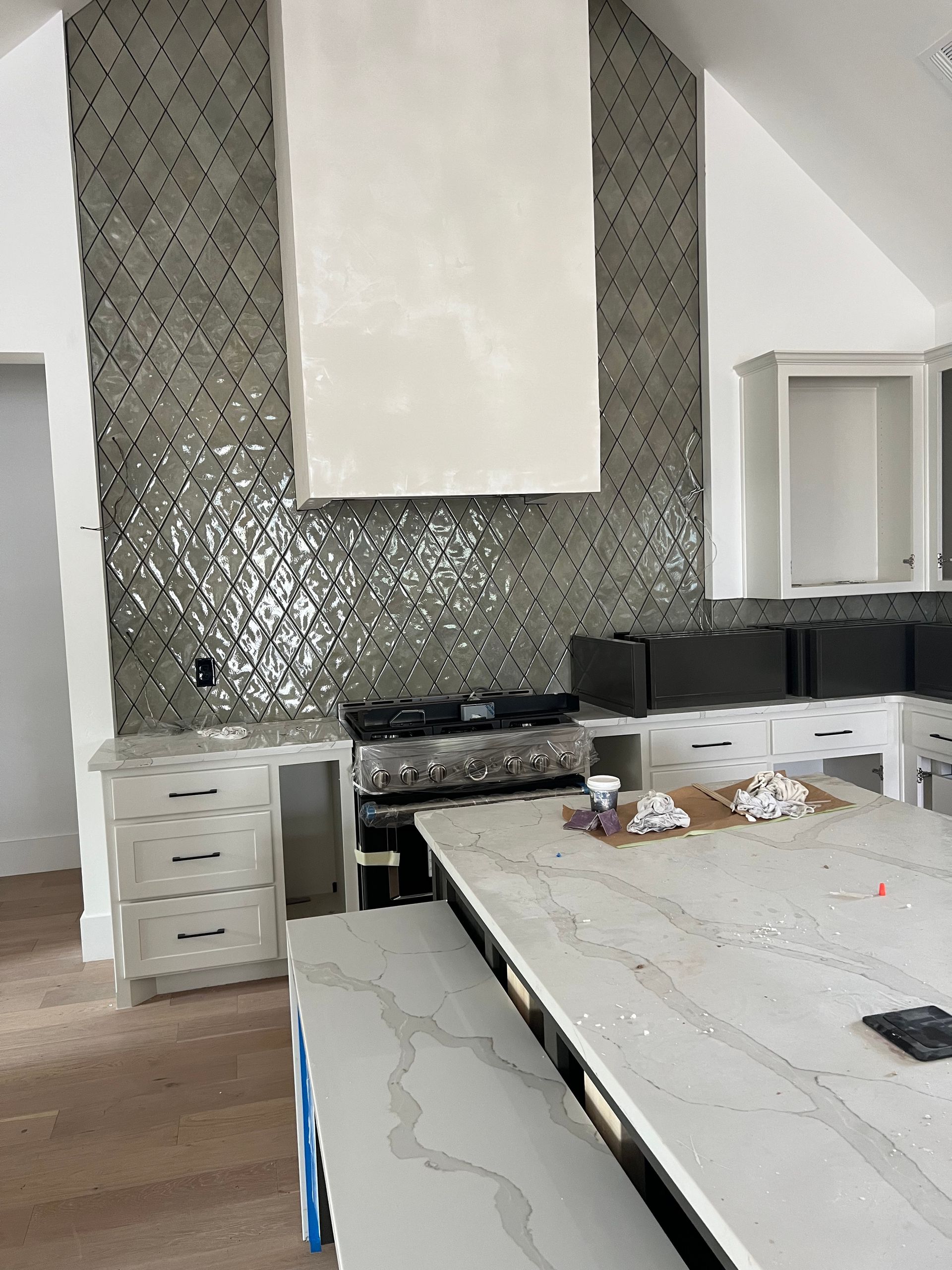 Kitchen under construction: stove with white range hood and mosaic tile backsplash, island with quartz countertop.