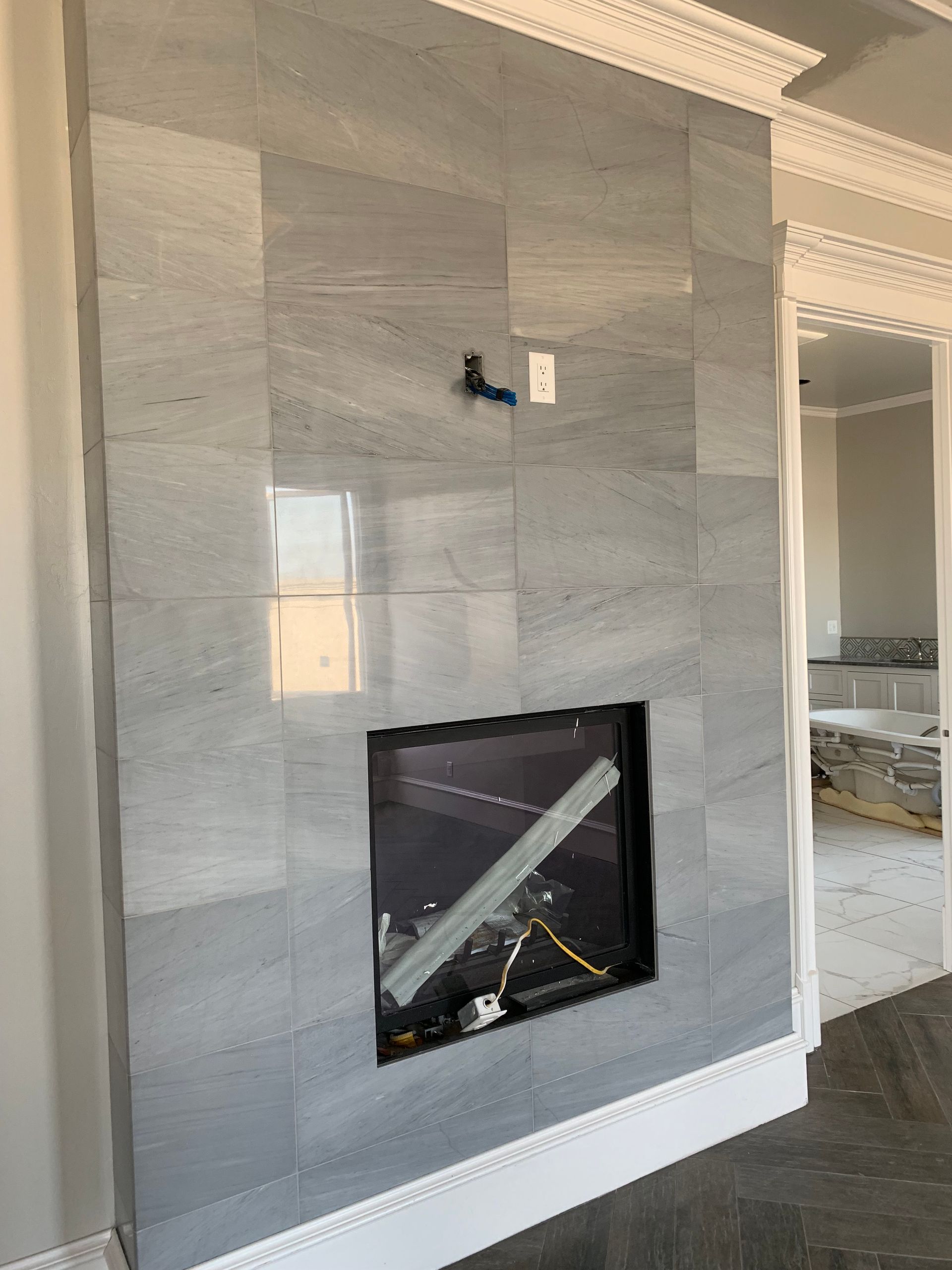 Tiled fireplace with a rectangular opening. Gray and white tiles. Electrical wires and a bracket are visible.