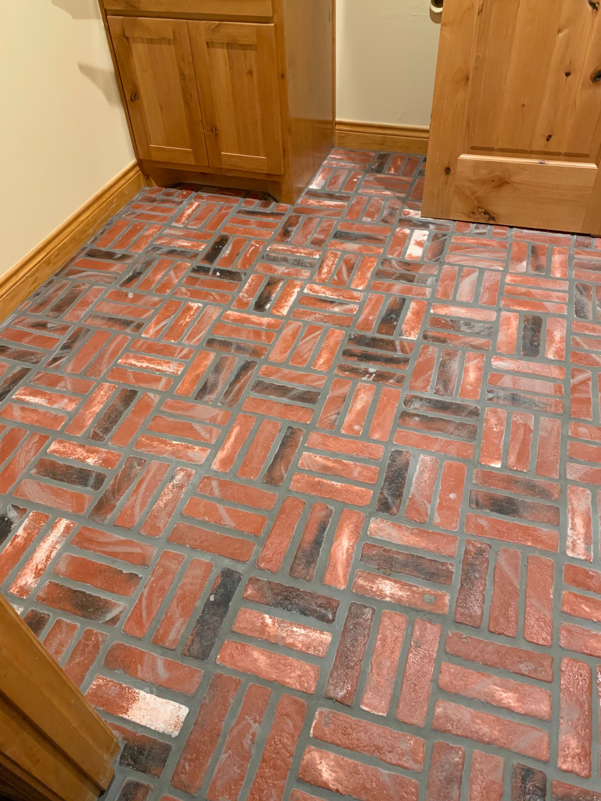 Brick flooring in herringbone pattern, red and black bricks with gray grout, inside a room with wooden cabinets and a door.