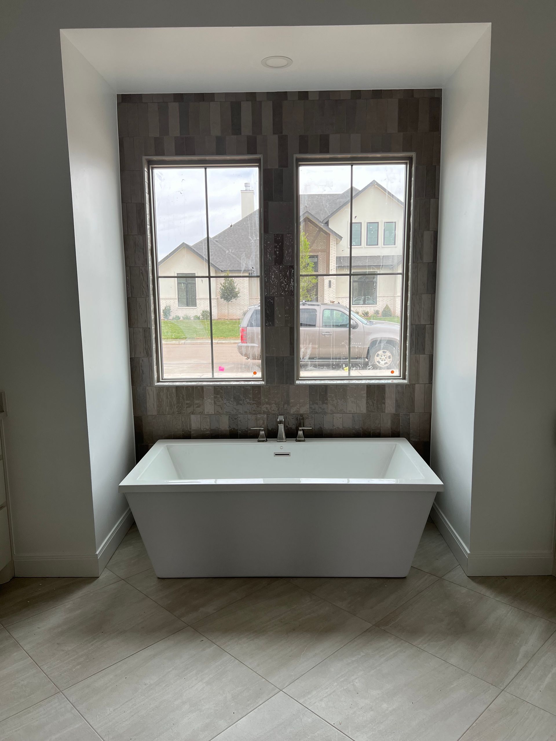 Freestanding white bathtub under two windows with gray tile backdrop.