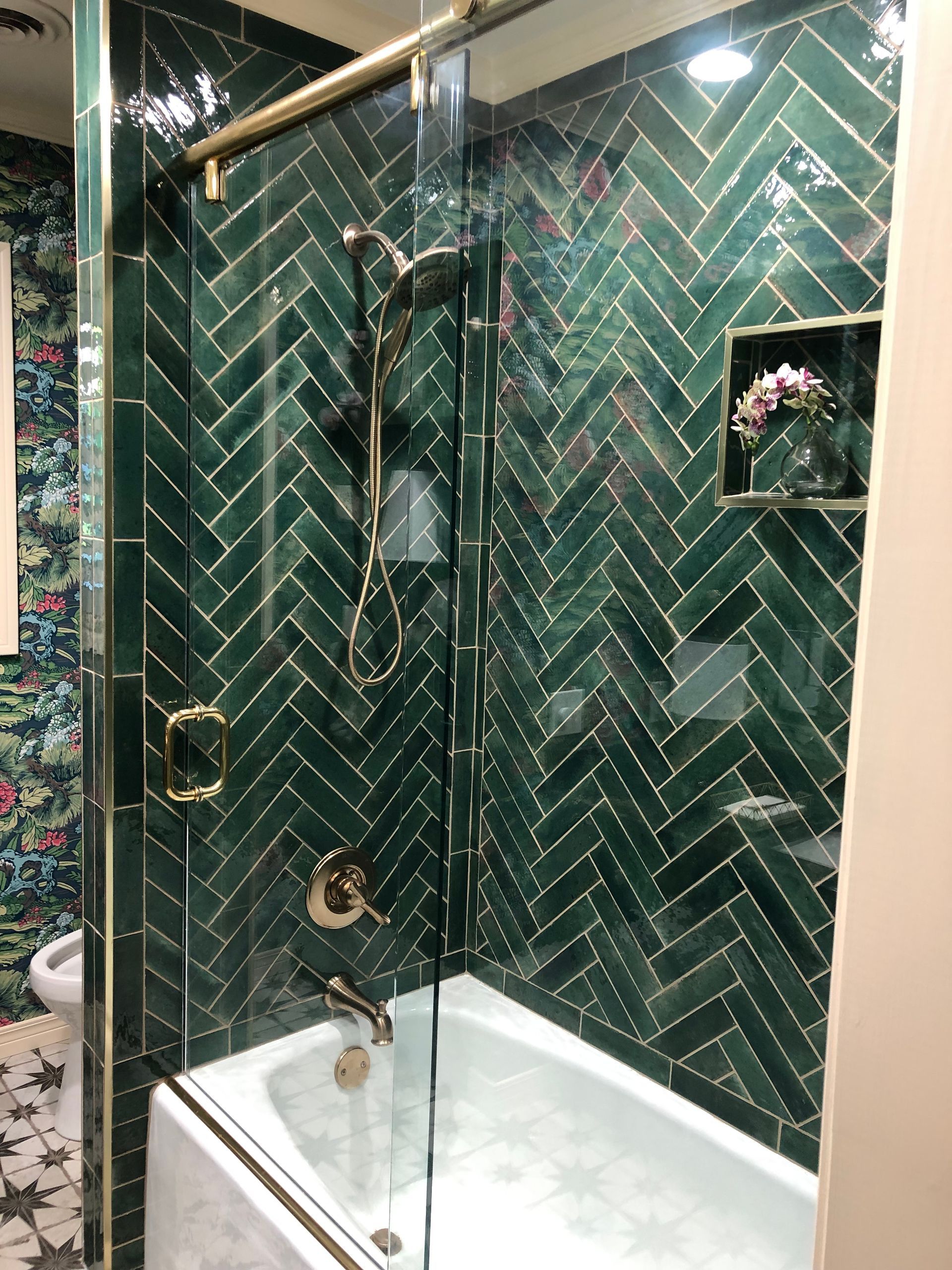 Emerald green herringbone-tiled shower with gold fixtures, a built-in shelf with flowers, and a glass door.