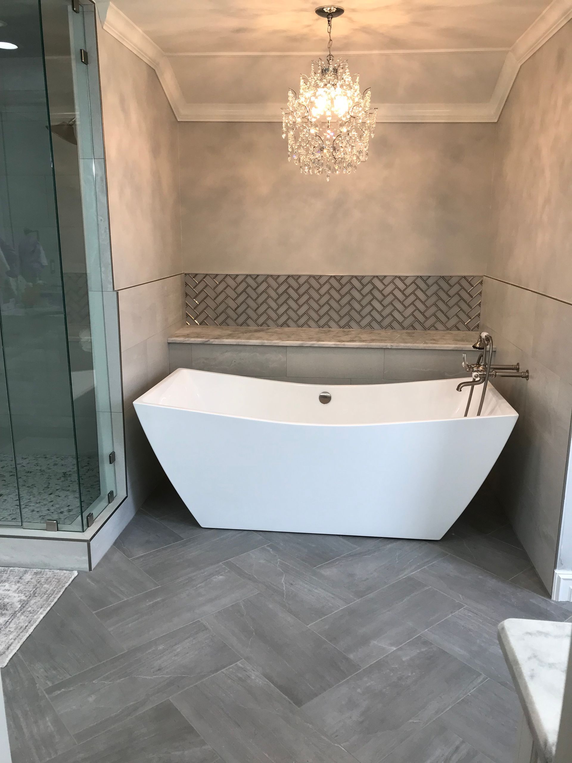 Luxurious white bathtub in a bathroom, gray herringbone tile floor, accent tile, chandelier.