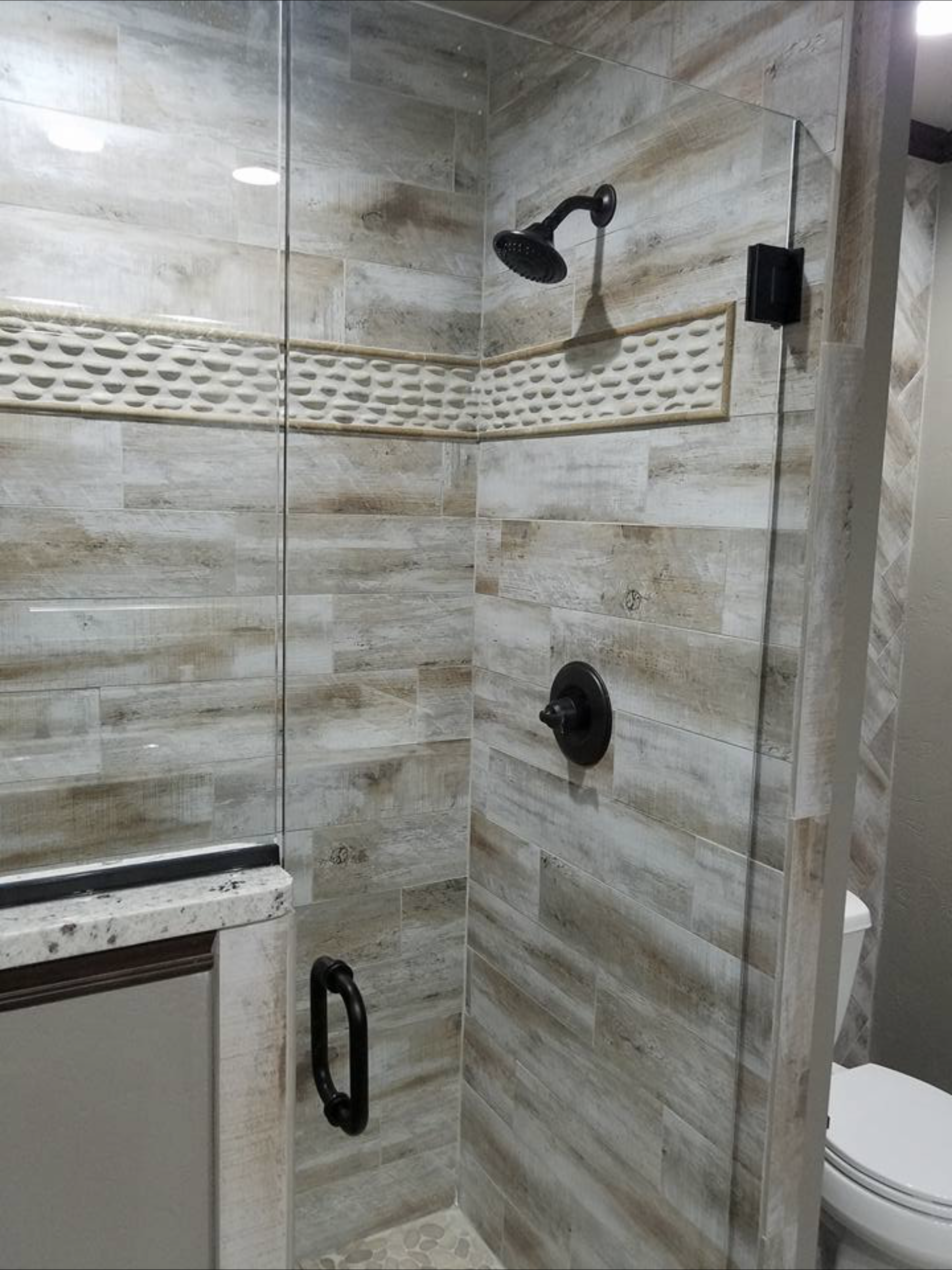Bathroom shower with glass door and brown, textured tiles, dark shower head and handle.