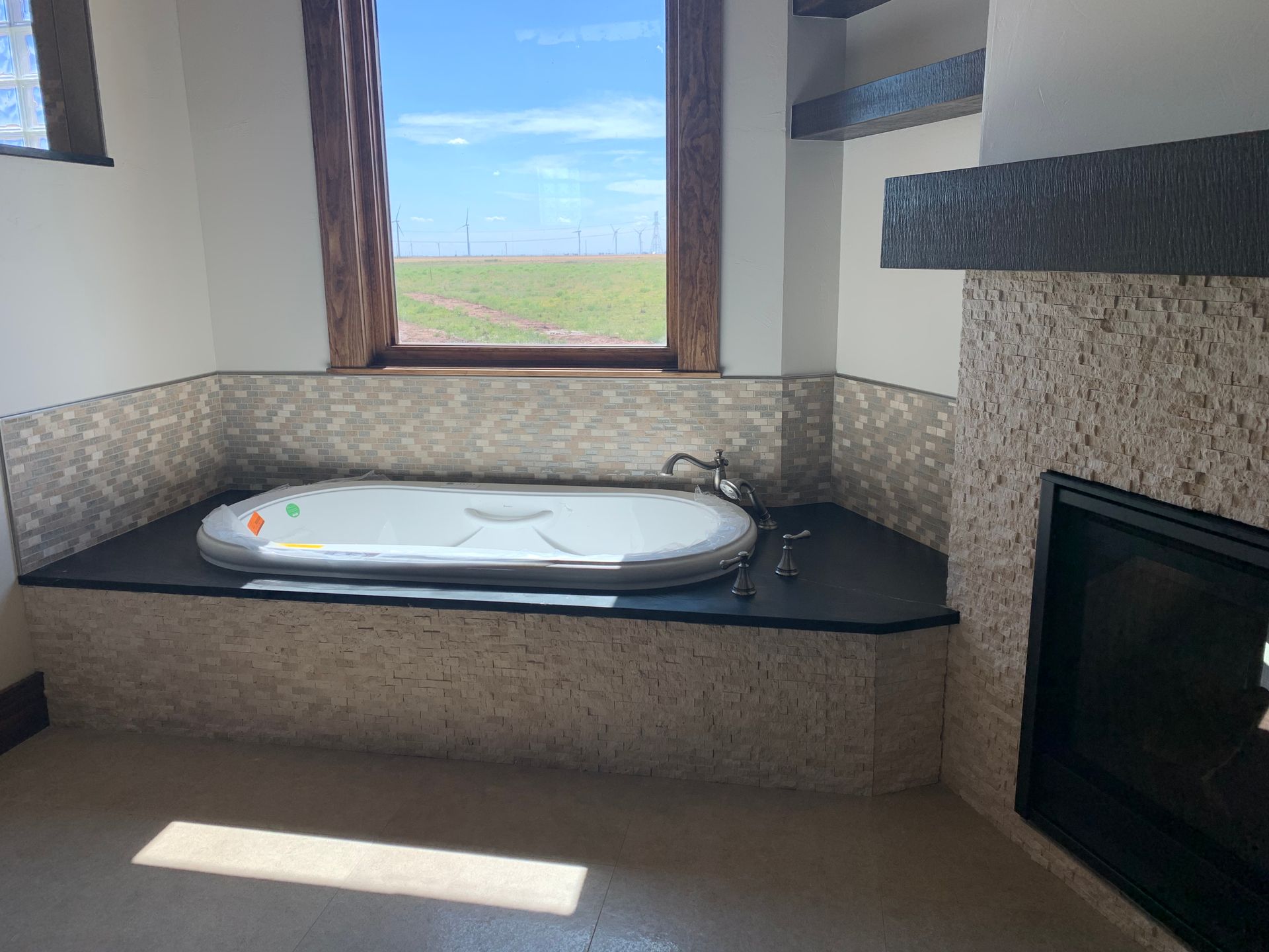 Bathroom with oval-shaped tub in a stone surround, next to a fireplace, with a window overlooking a field.