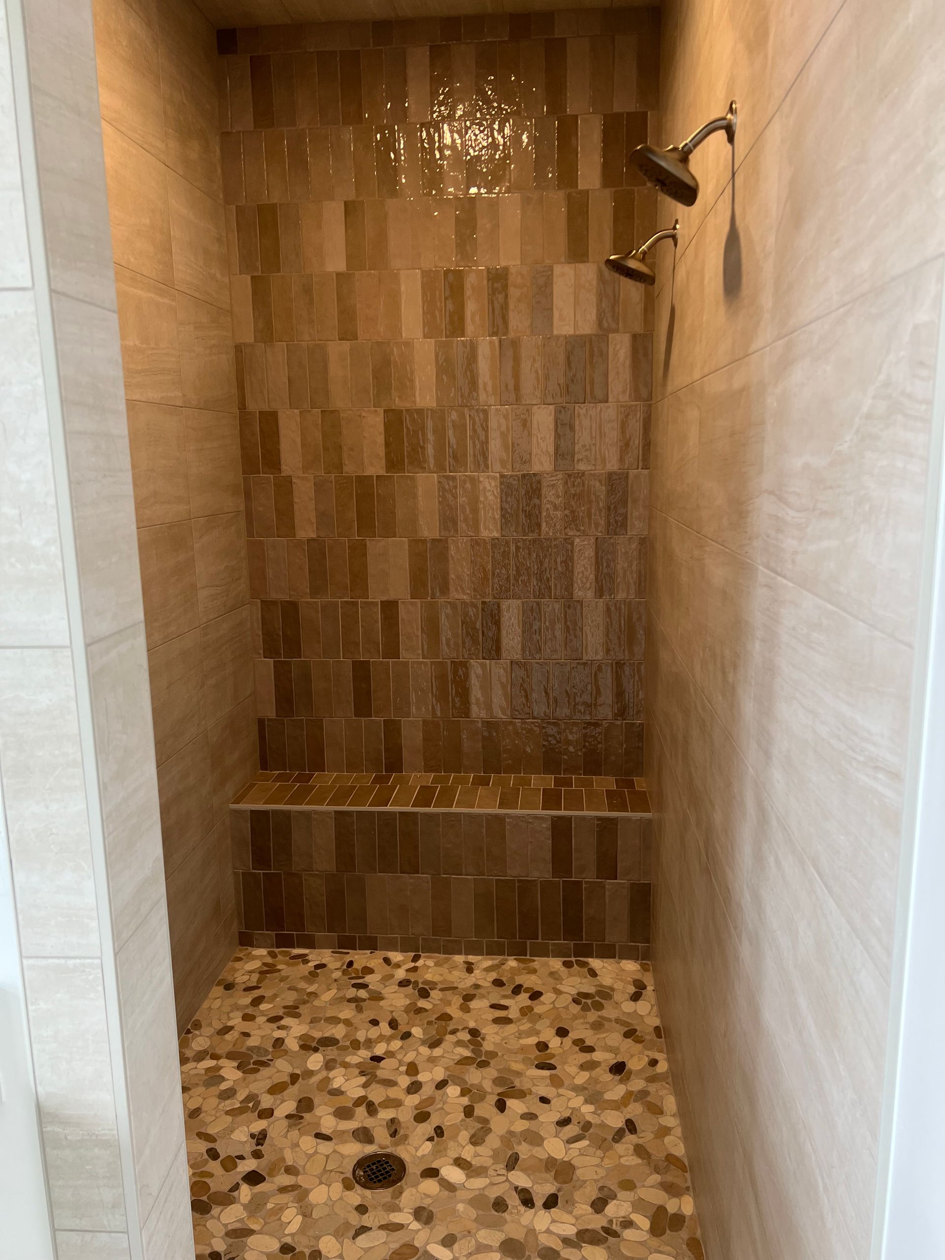 Shower with pebble floor, tiled walls in shades of brown, and two showerheads. Includes a built-in bench.