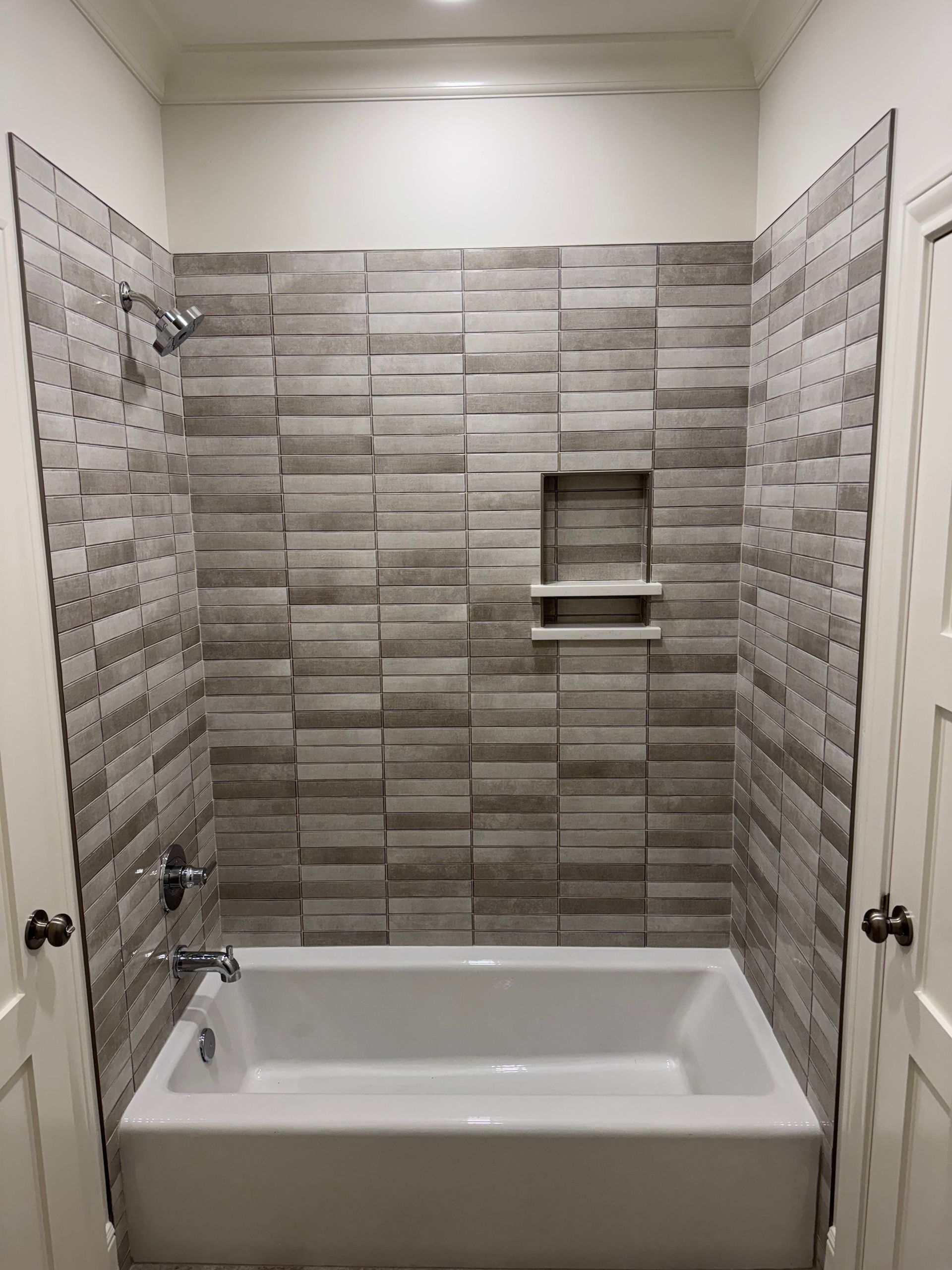 Bathroom with white tub, grey tile walls, showerhead, and a built-in shelf.