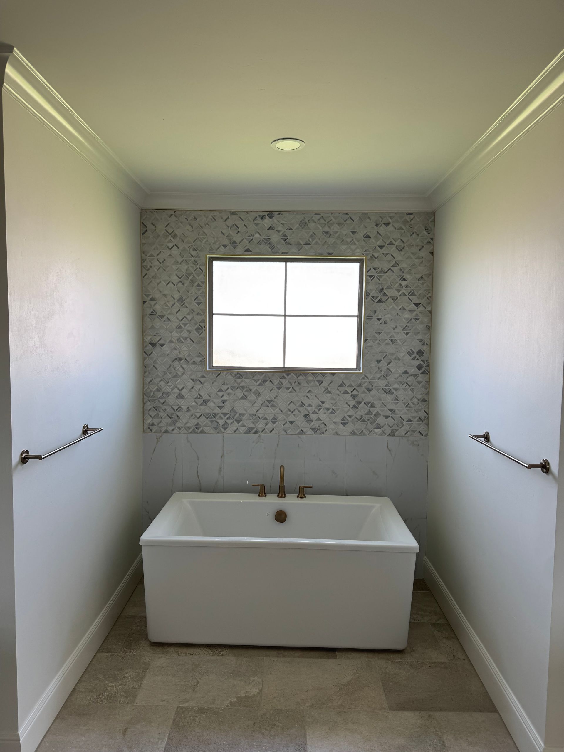A white bathtub centered under a window in a bright bathroom with neutral walls and decorative towel racks.