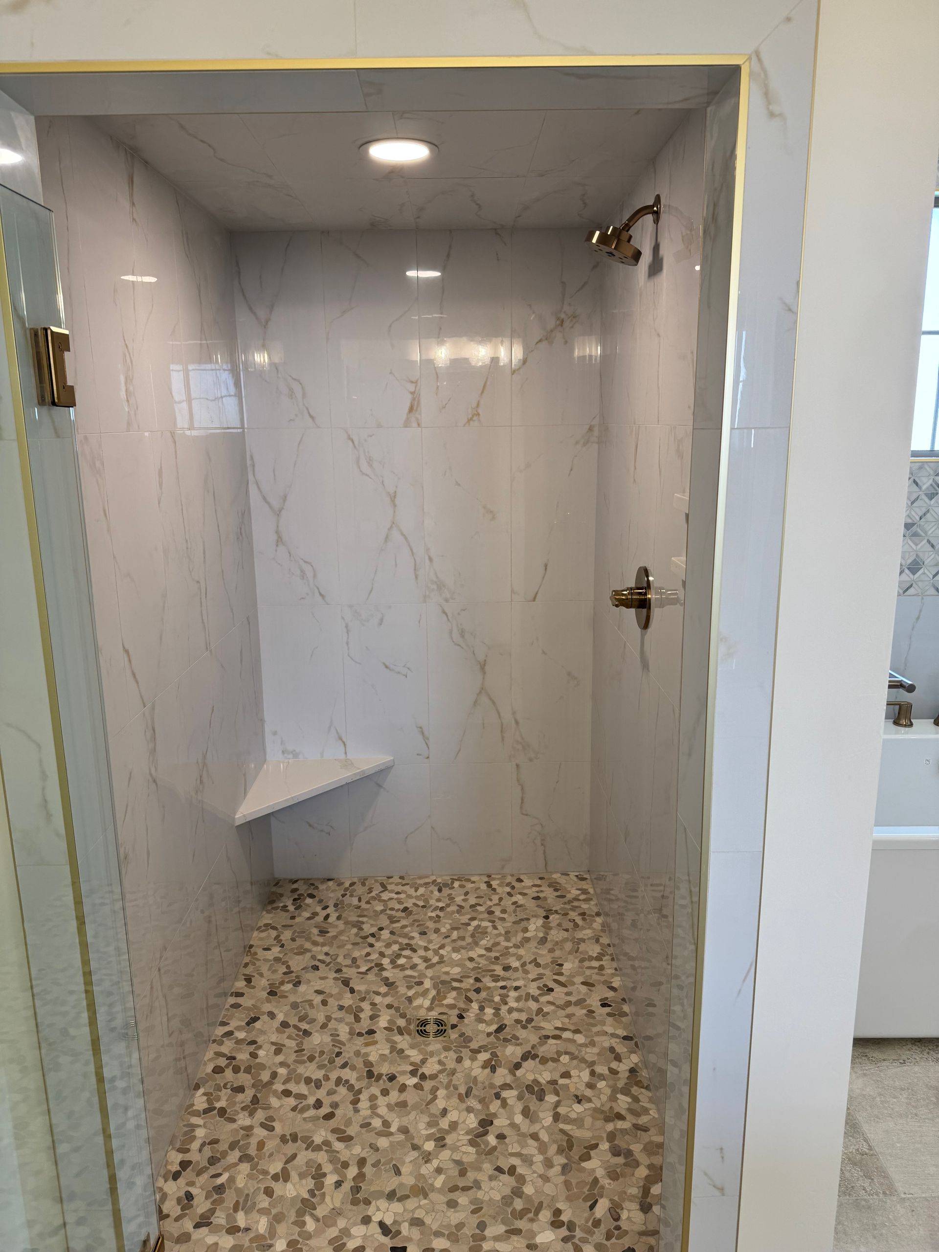 Shower with white marble walls, pebble floor, built-in seat, and gold fixtures.