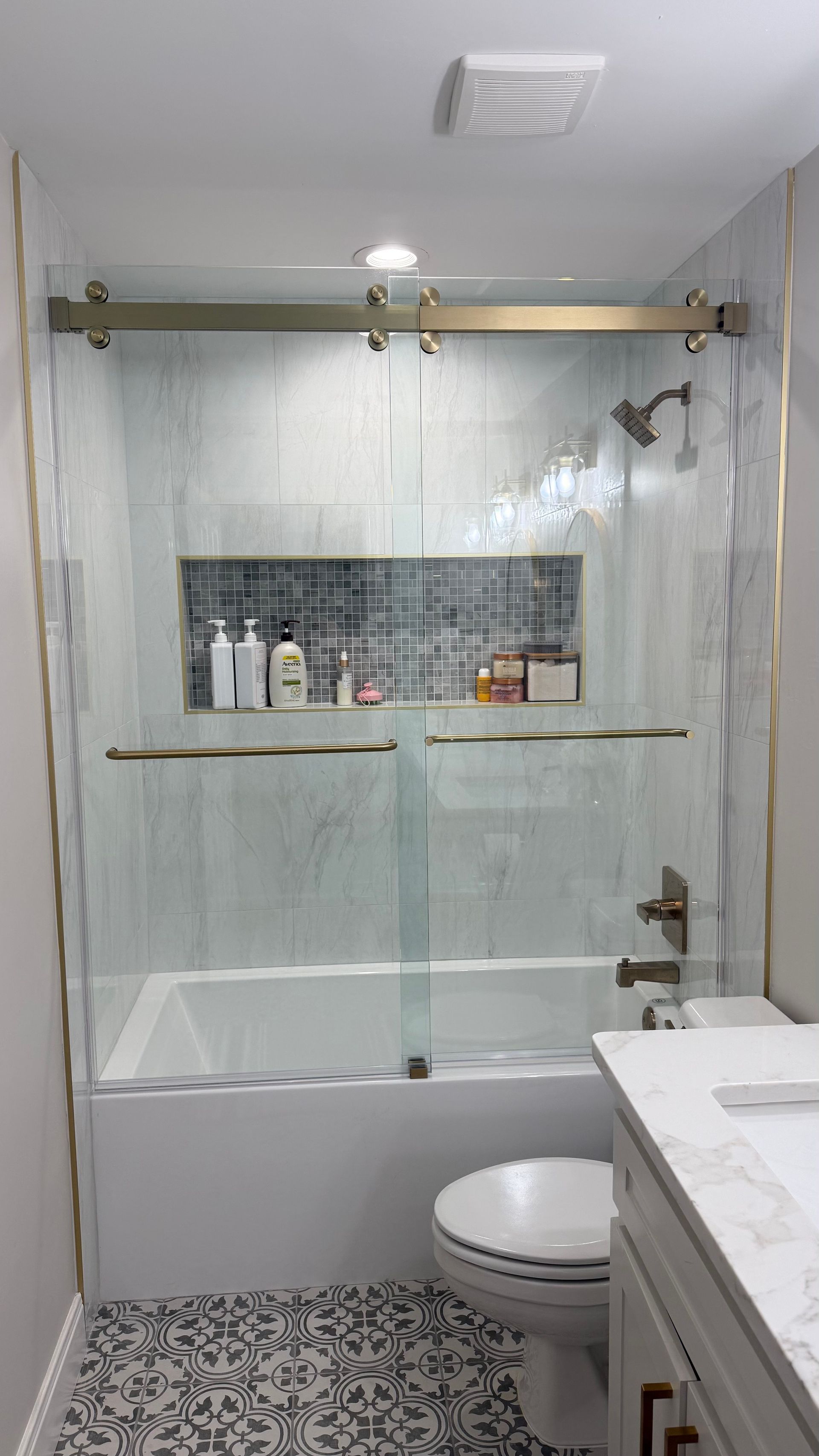 Bathroom with a glass shower door, gold trim, and a built-in shelf. White tub and toilet.