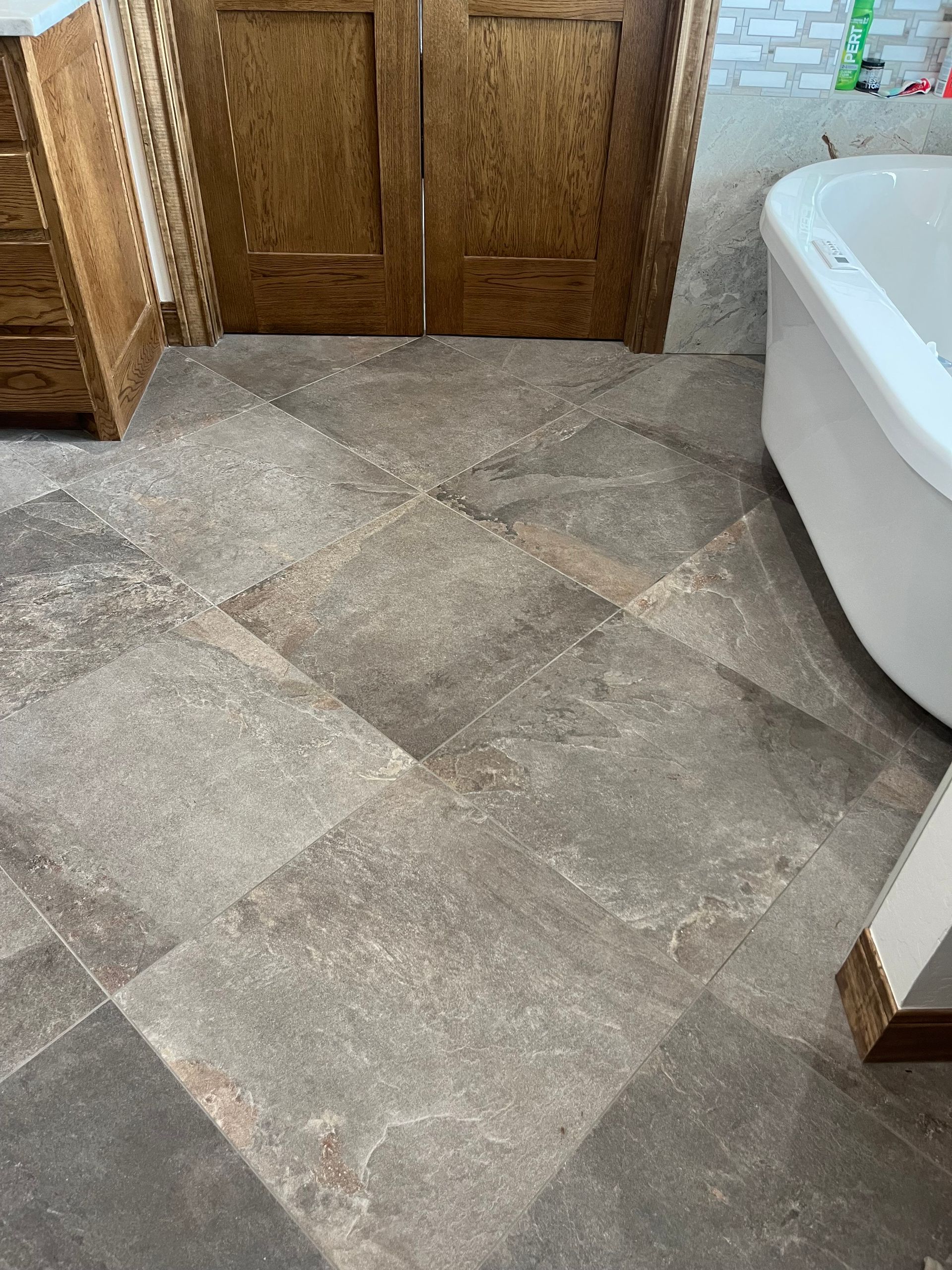 Bathroom floor with large, gray-brown tiles laid in a diagonal pattern; wooden doors and white bathtub in view. Bathroom floor with large, gray-brown tiles laid in a diagonal pattern; wooden doors and white bathtub in view.