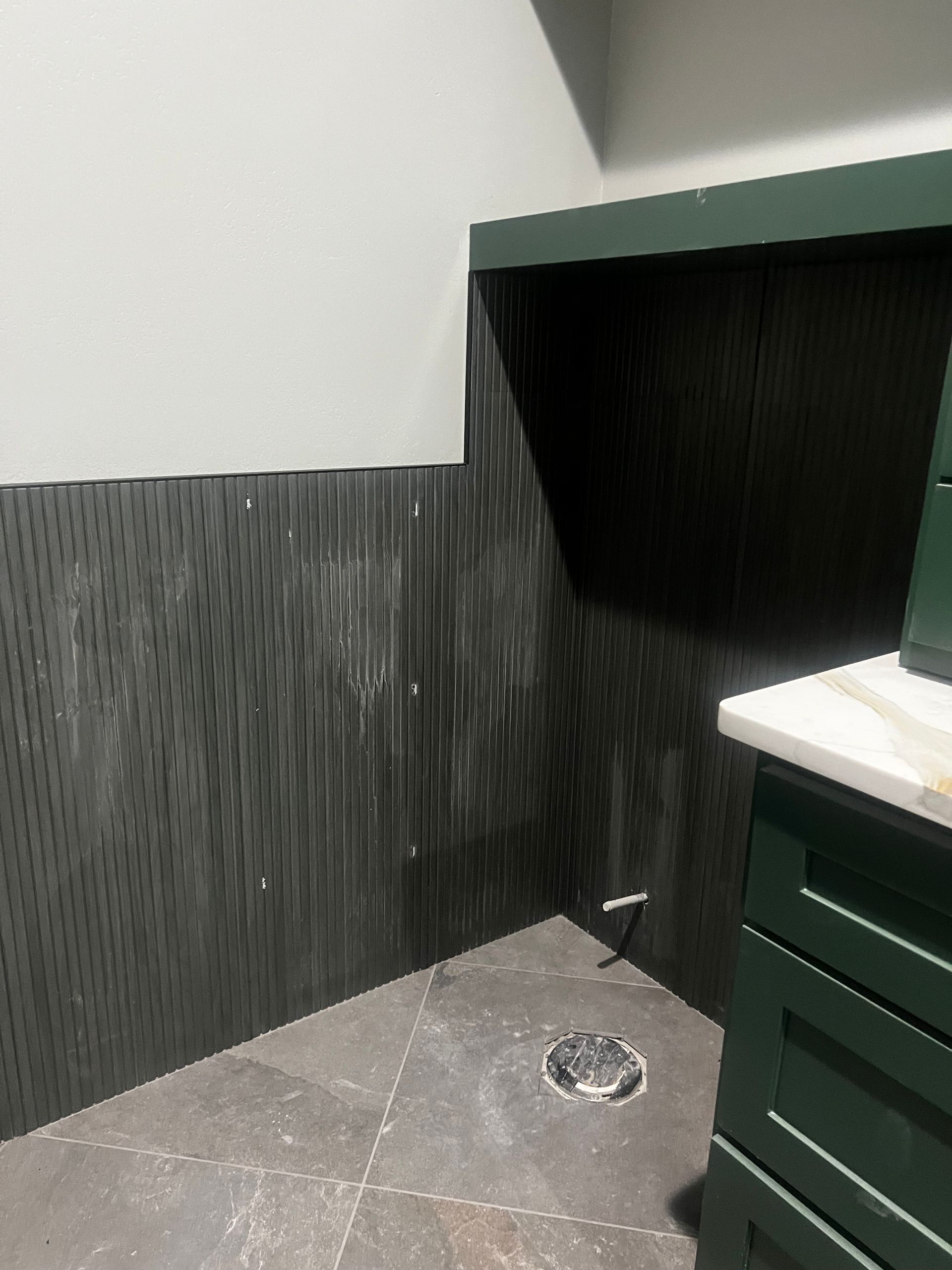 Dark green-tiled wall in a bathroom with dark cabinets, a white countertop, and gray tiled floor.