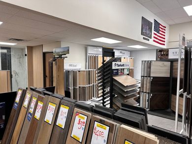 A flooring showroom displays samples of wood and tile flooring. American flag hangs on the wall.