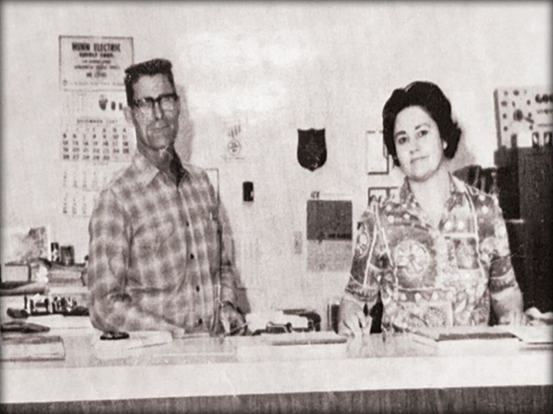 Man and woman behind a counter. Man wears glasses and plaid shirt. Woman in printed blouse. Office setting, calendar visible. Man and woman behind a counter. Man wears glasses and plaid shirt. Woman in printed blouse. Office setting, calendar visible.