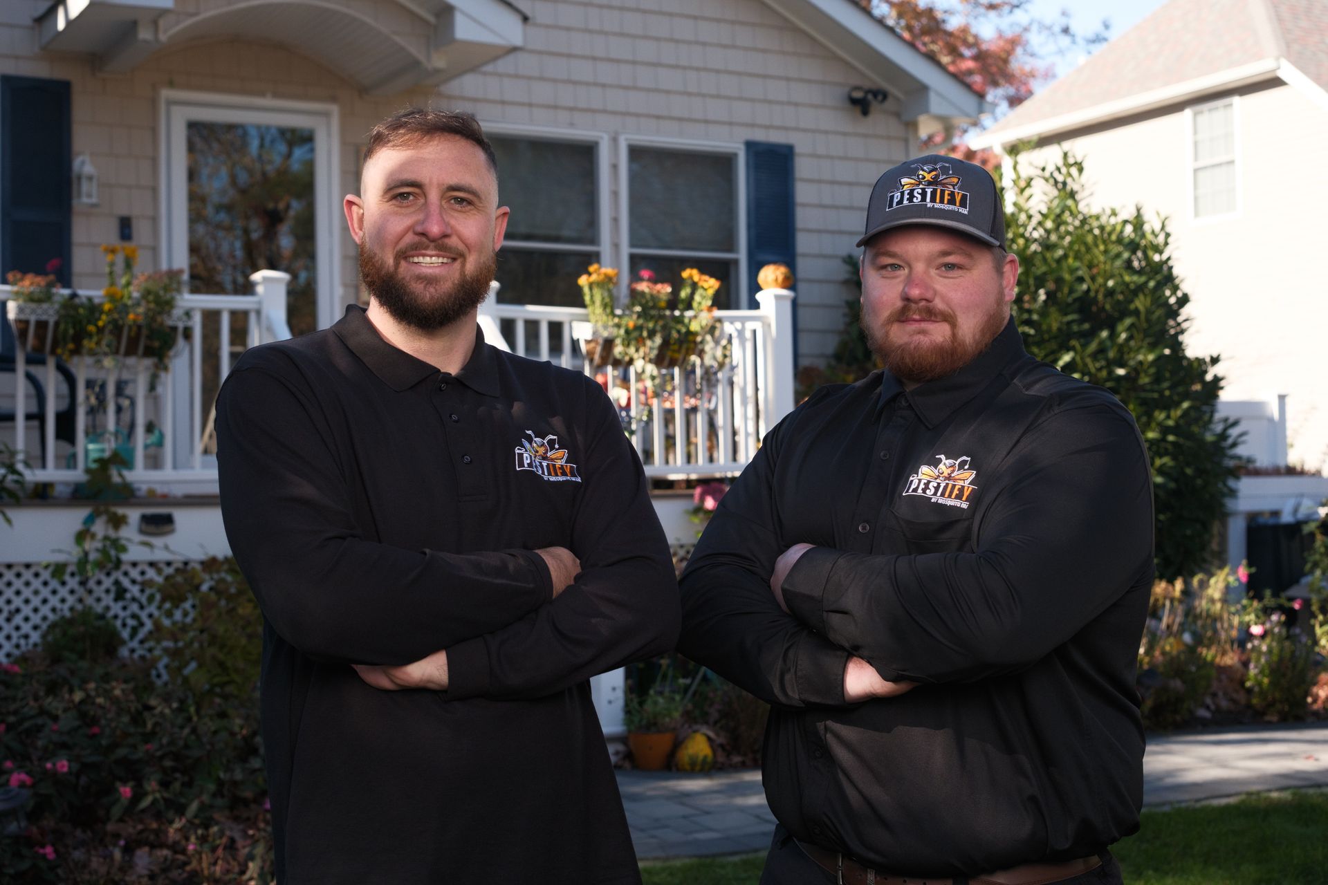 Two professionals in matching black uniform shirts stand with arms crossed in front of a house with a white porch.