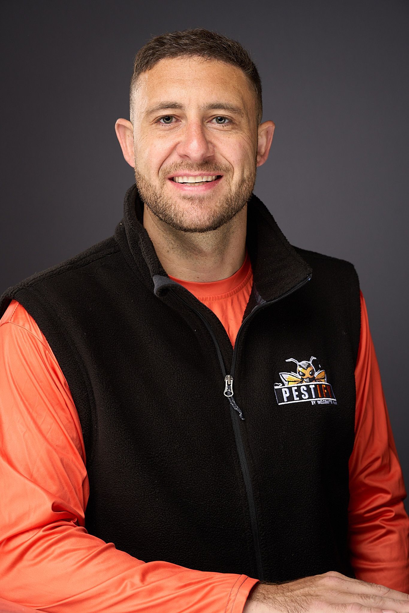 A person with a beard wearing an orange long-sleeve shirt and cap stands with arms crossed in front of a house.