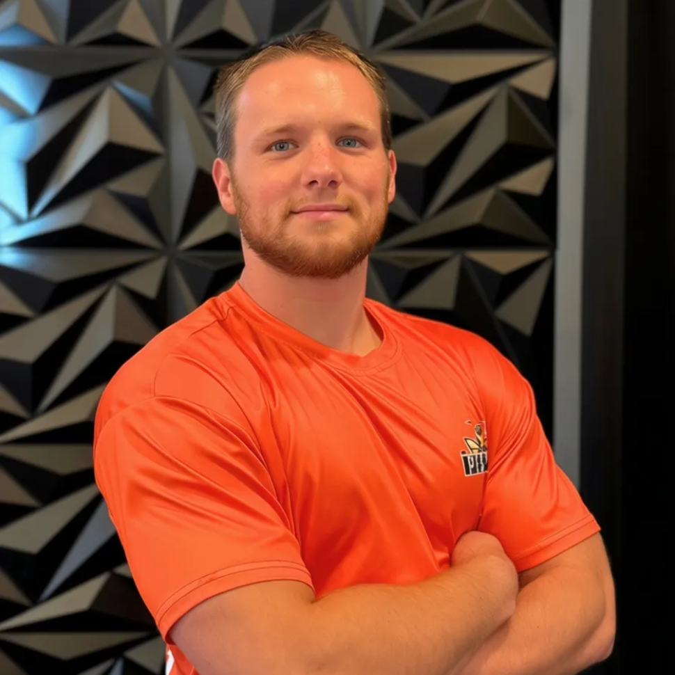 A man with a short beard wearing an orange t-shirt, standing with arms crossed in front of a black geometric wall.