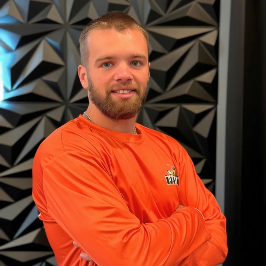 A smiling person with a beard, wearing an orange long-sleeved shirt with arms crossed, in front of a geometric wall.