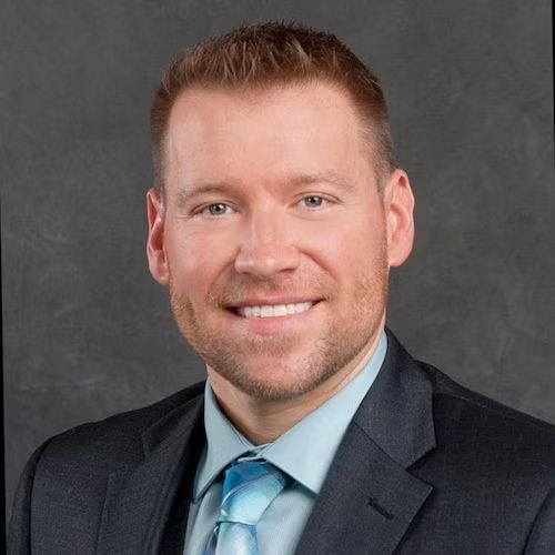 Professional headshot of a person wearing a dark suit, light blue dress shirt, and a bright blue tie against a gray backdrop.