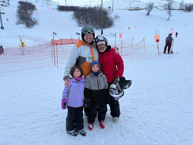 A family of four wearing winter gear and ski helmets stands together on a snowy ski slope.