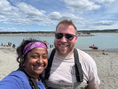 A smiling couple poses for a selfie on a sunny, sandy beach by a lake with people and a red boat in the background.