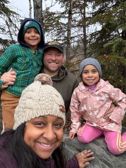A group of four smiling people outdoors in a wooded area, wearing winter hats and jackets.