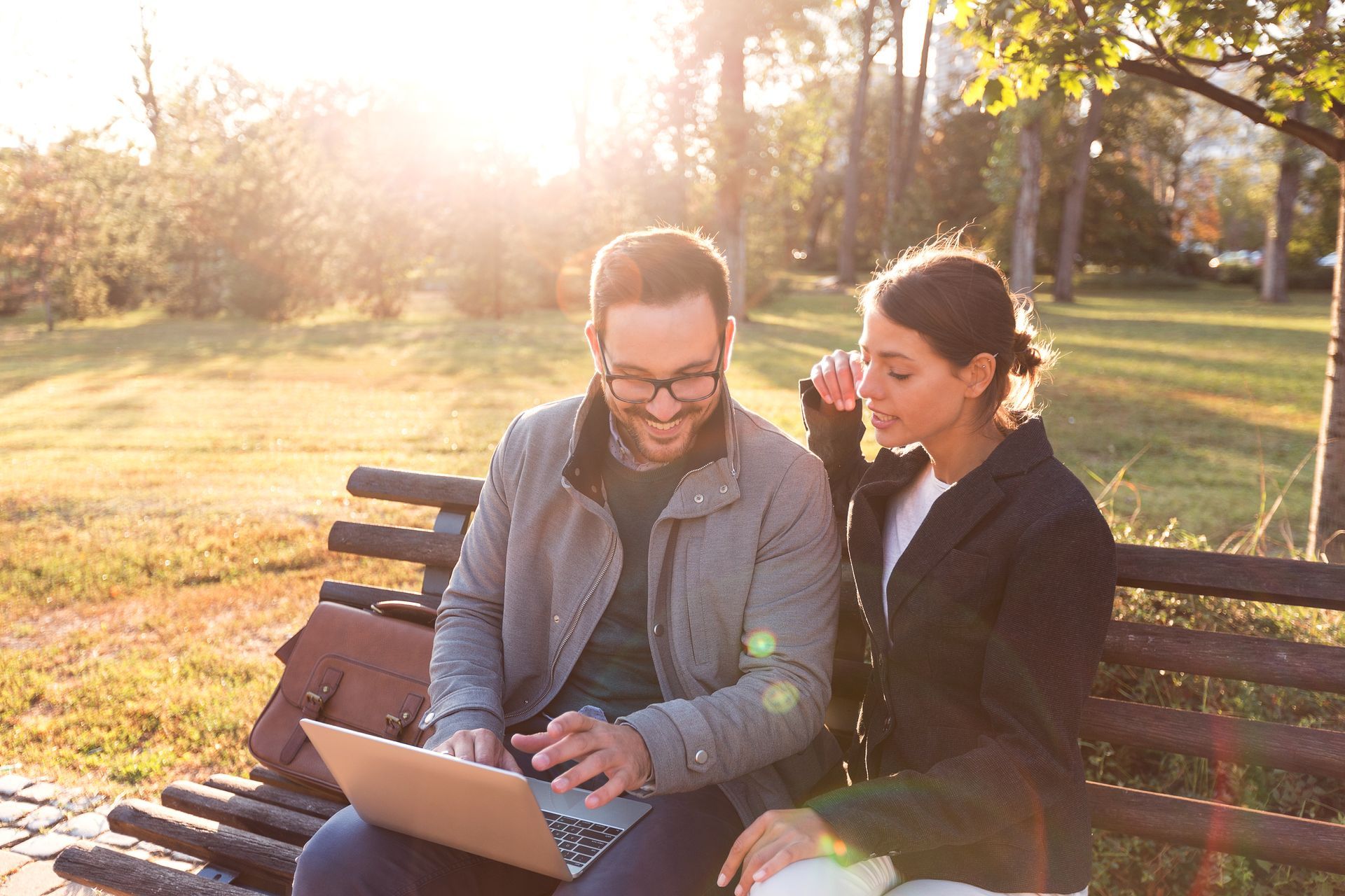 Two people sit together on a park bench, looking at a laptop computer during a bright, sunny afternoon.