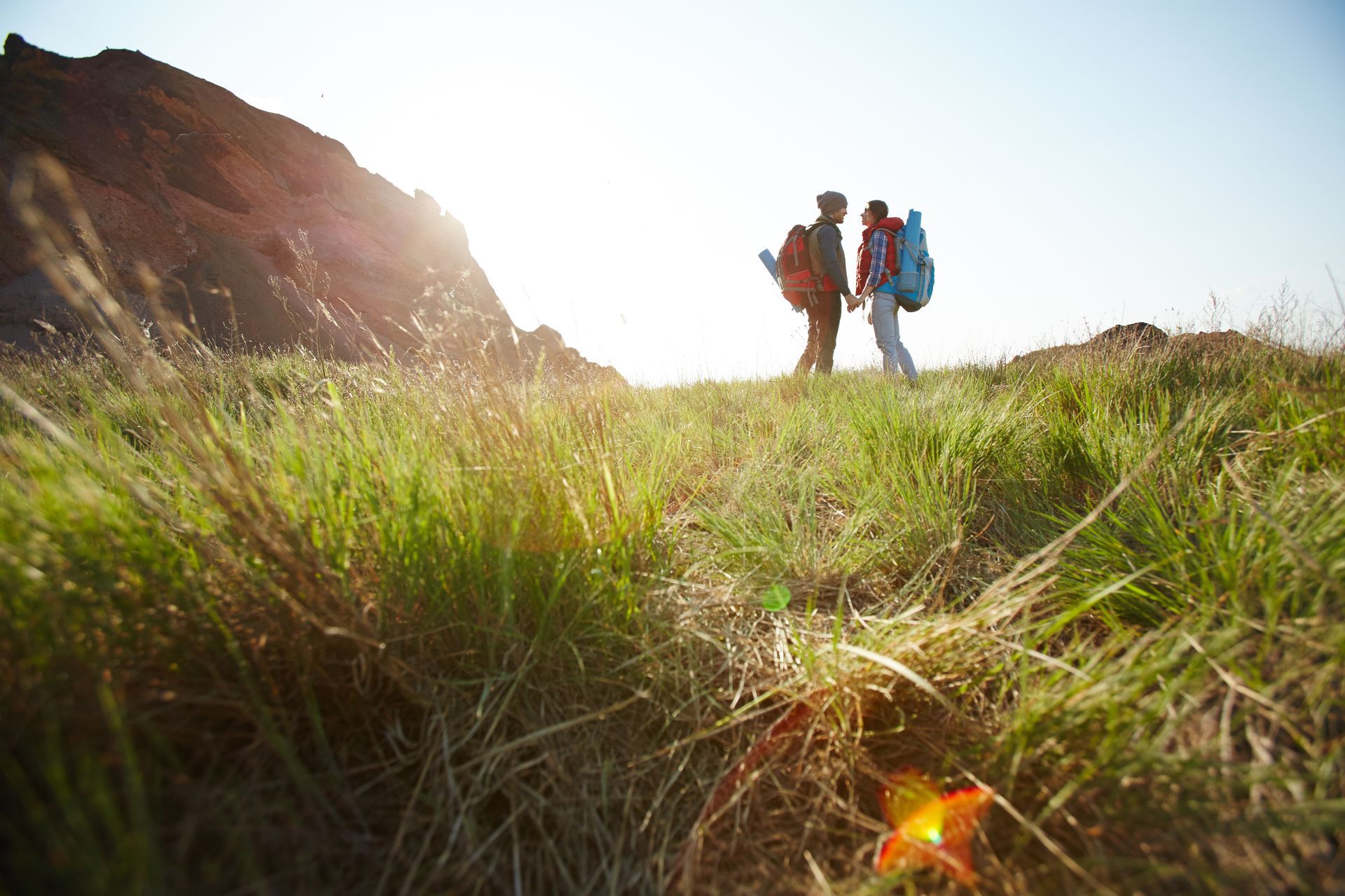 Two backpackers stand in a grassy field facing each other against a bright, sunlit mountain backdrop.