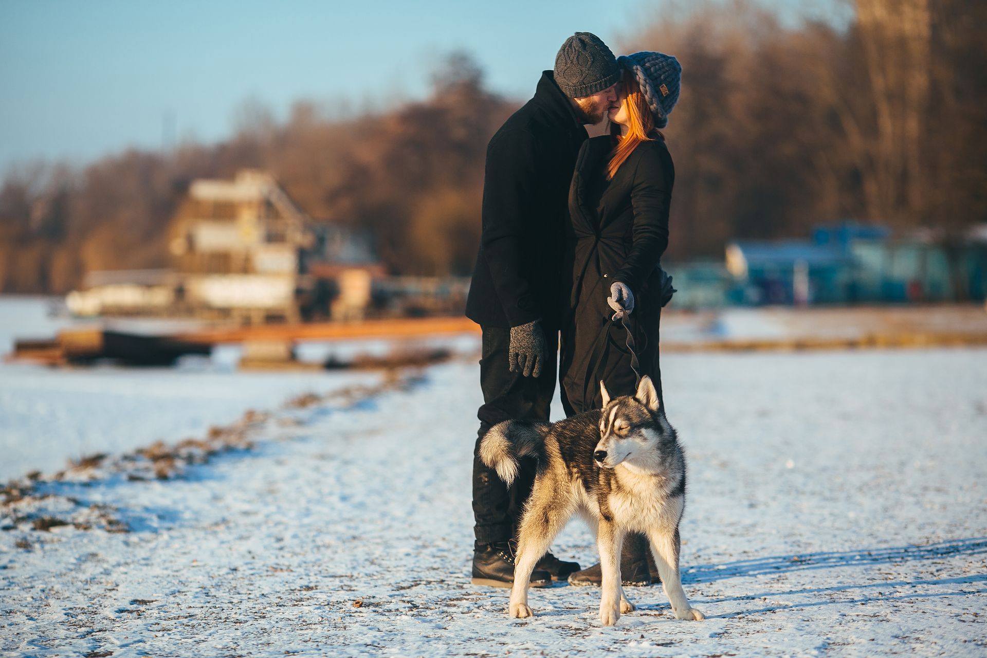 A couple in dark winter clothing kisses on a snowy beach while holding the leash of a standing husky.