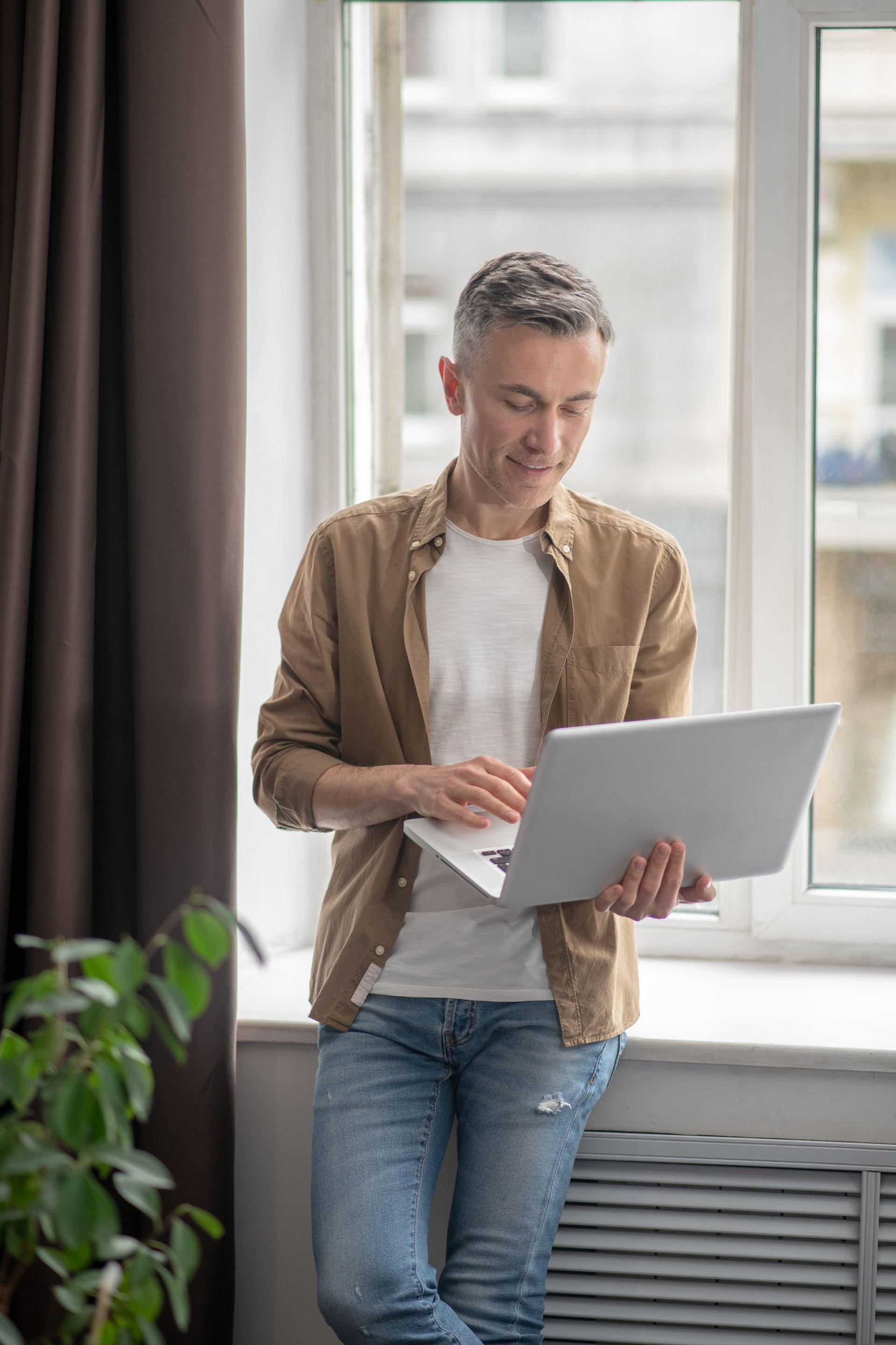 A person wearing a tan shirt and jeans stands by a window, looking down at a laptop held in their hands.