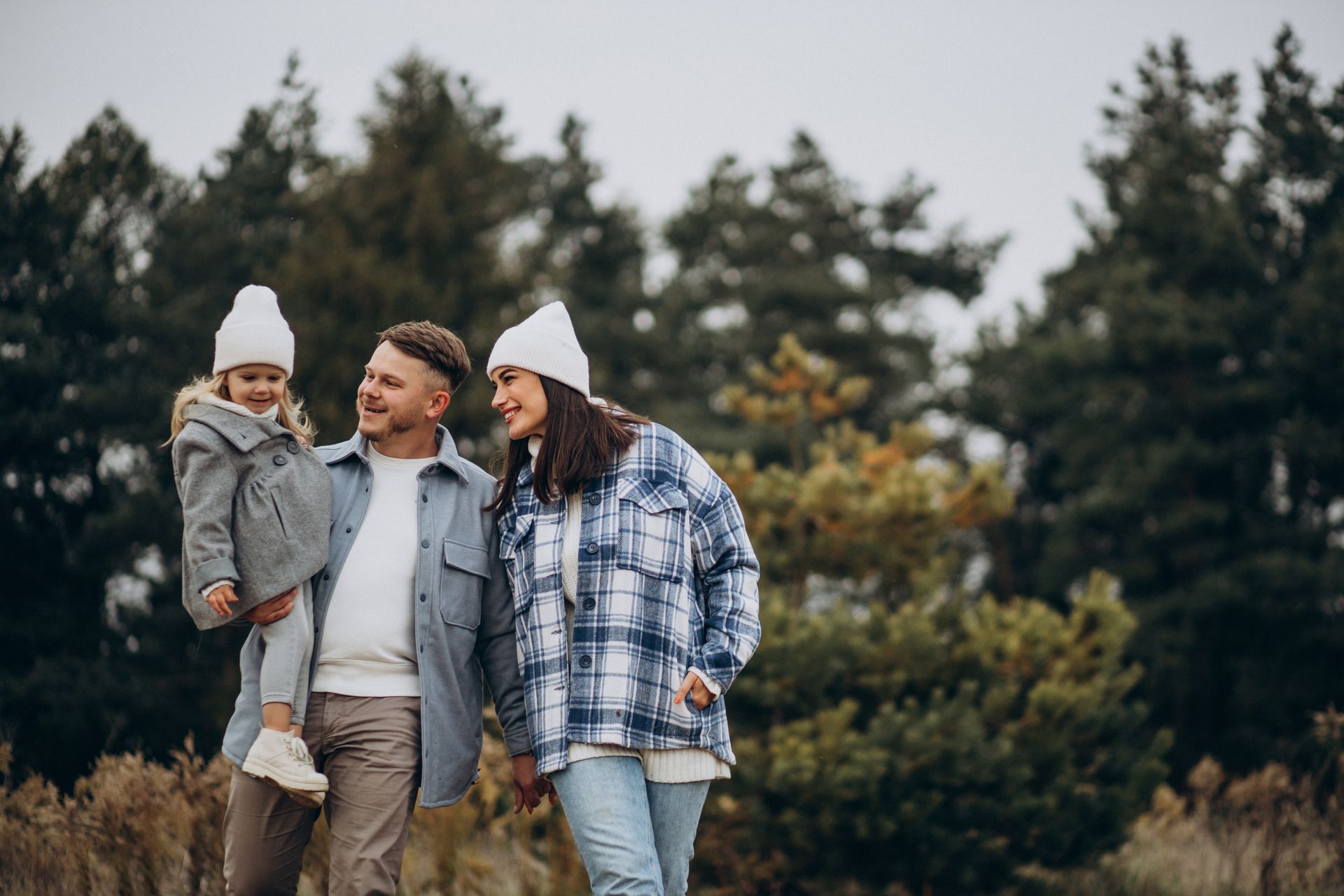 A family of three wearing white beanies and warm layers, walking outdoors in a natural setting with evergreen trees.