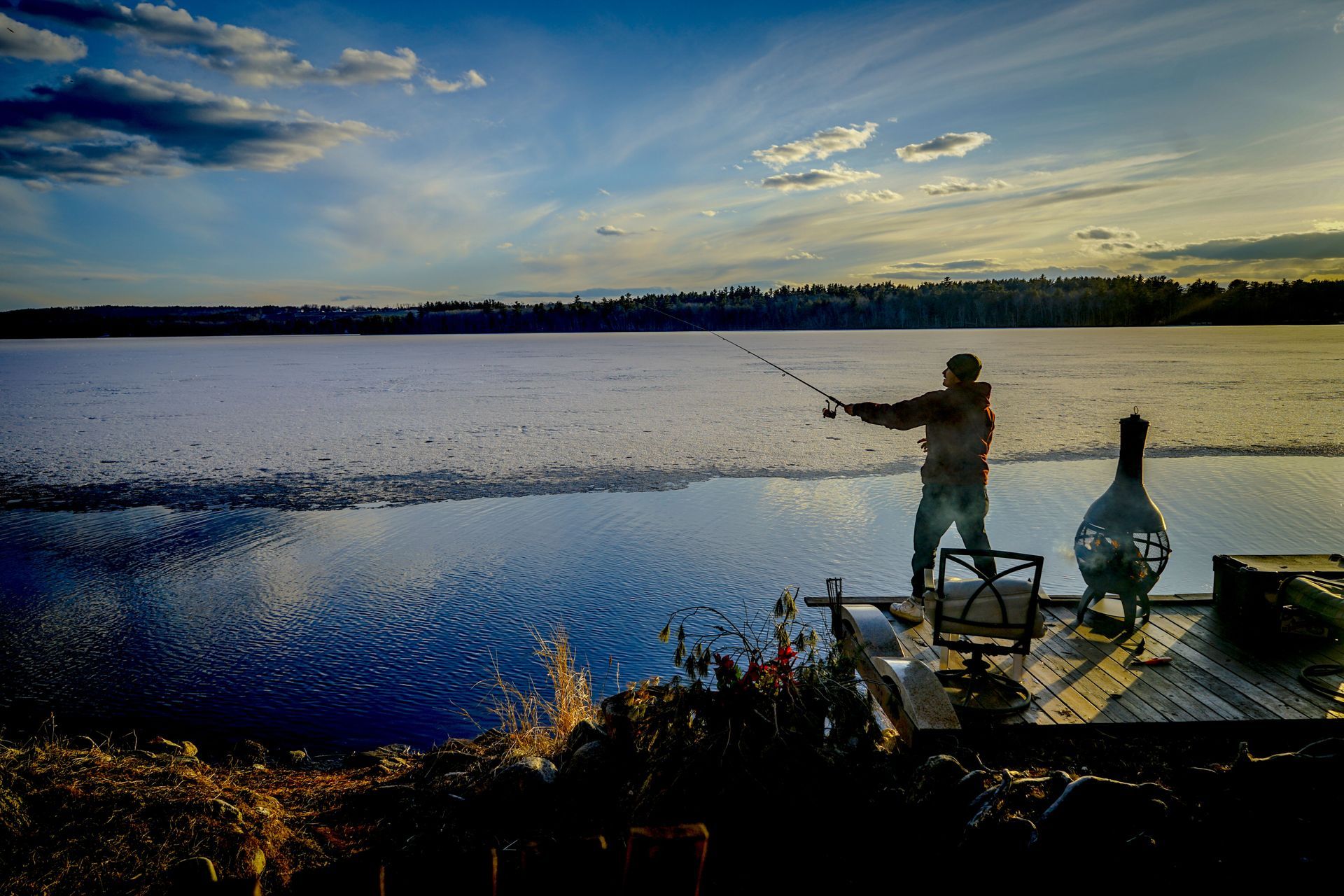 A silhouetted person casts a fishing line from a wooden dock into a large lake at sunset, with ice covering the far water.