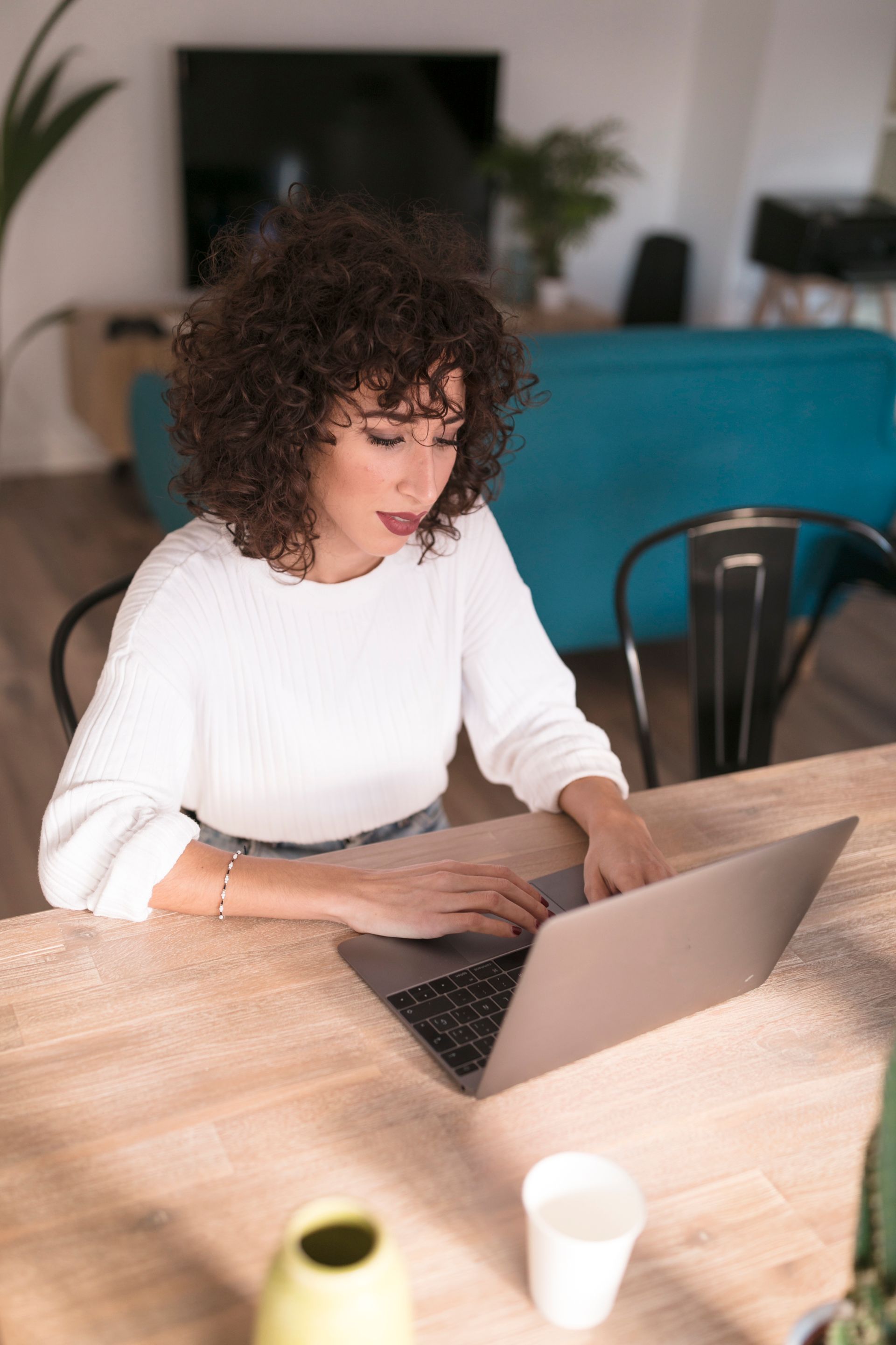 A person with curly hair wearing a white long-sleeve shirt types on a laptop at a wooden table in a living room.