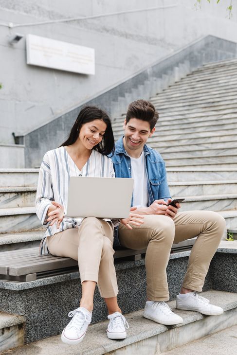 Two people sitting on outdoor stairs, one looking at a laptop and the other holding a smartphone, both smiling.