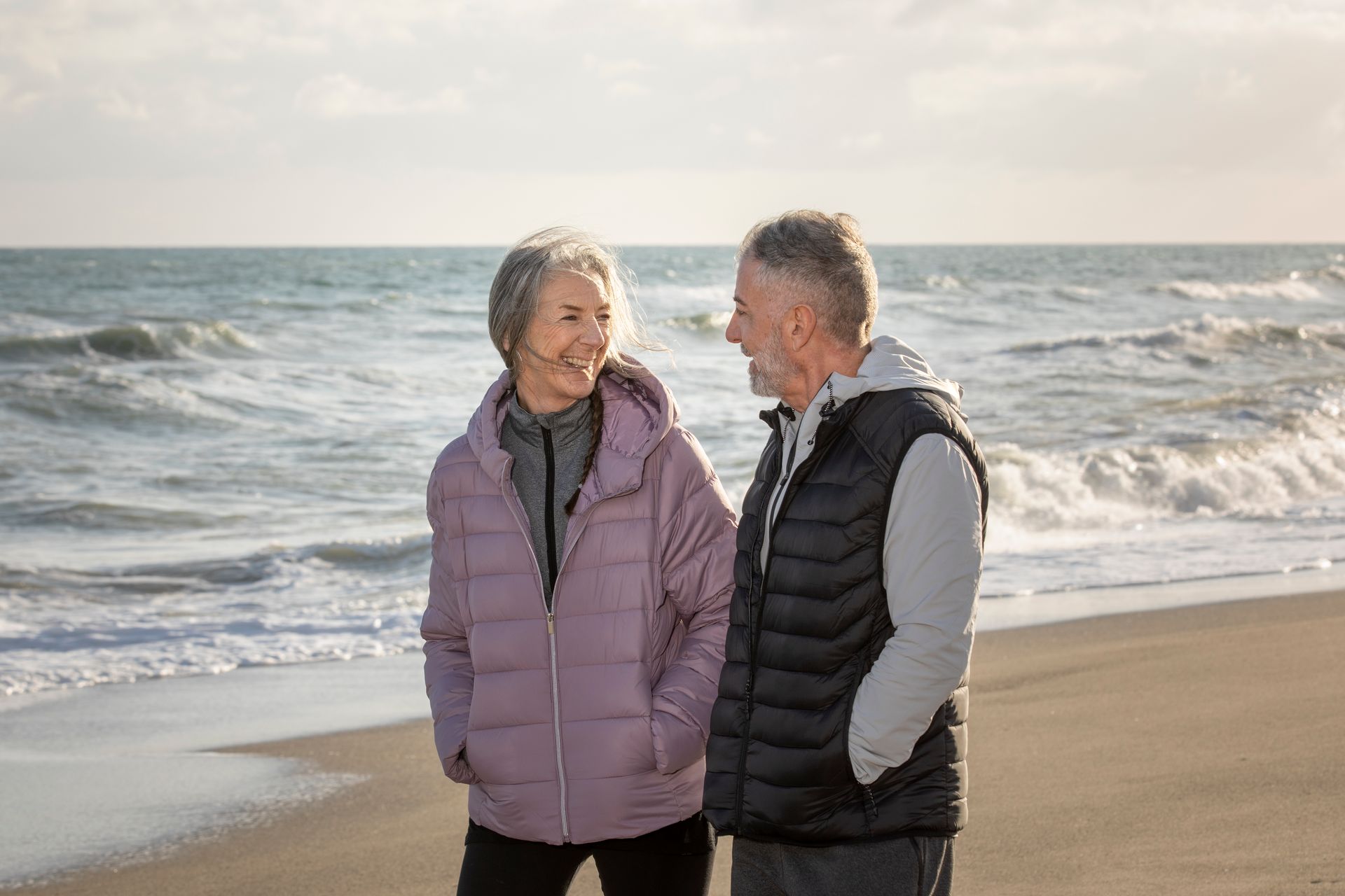 A couple in quilted jackets smiles at each other while walking along a sandy beach with crashing waves in the background.