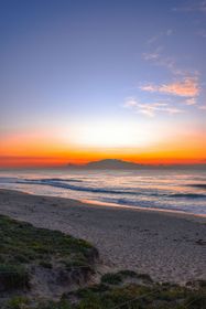 A vibrant sunset casts orange and blue hues over a sandy beach, with gentle waves rolling in toward a distant island.