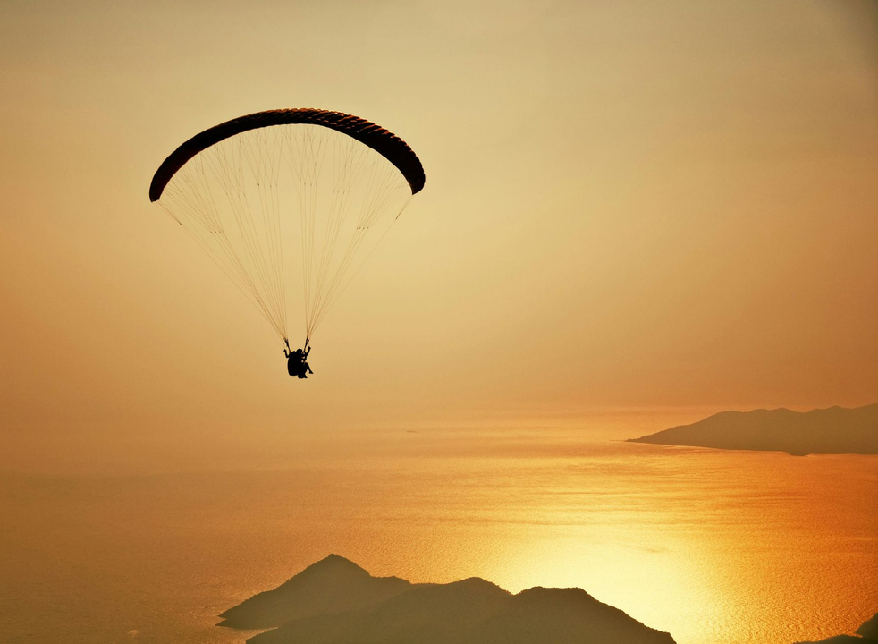 A person paragliding over a sunset ocean with mountainous coastline visible in the distance.