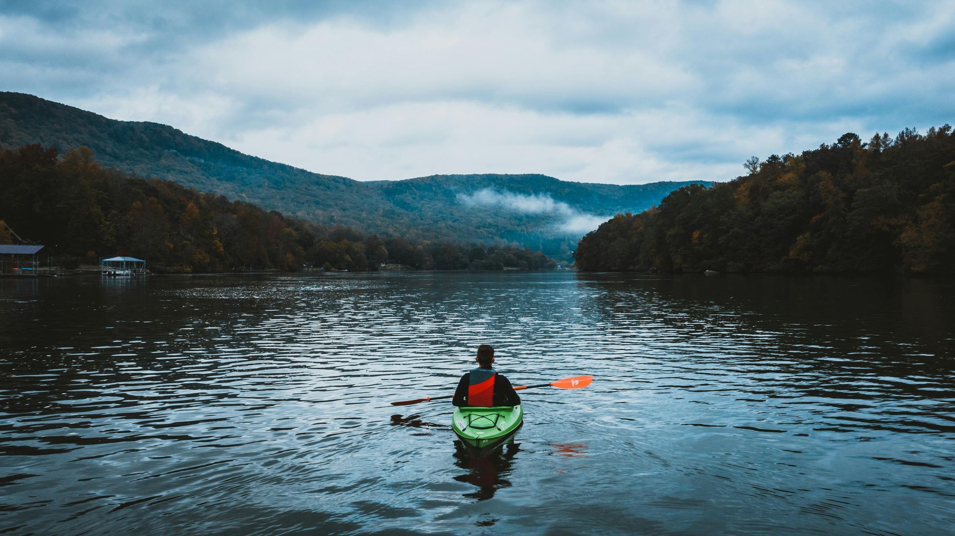A person paddles a green kayak on a calm, wide river between misty, forested mountains under a cloudy sky.