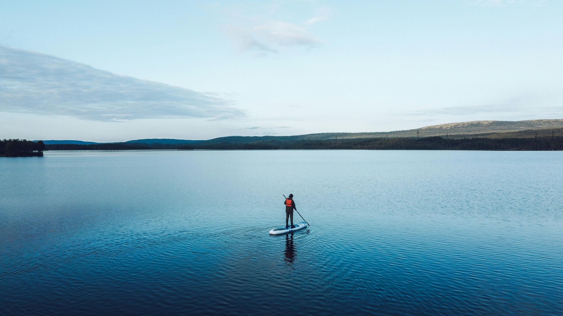 A person paddleboarding on a vast, calm blue lake surrounded by distant rolling hills under a soft sky.