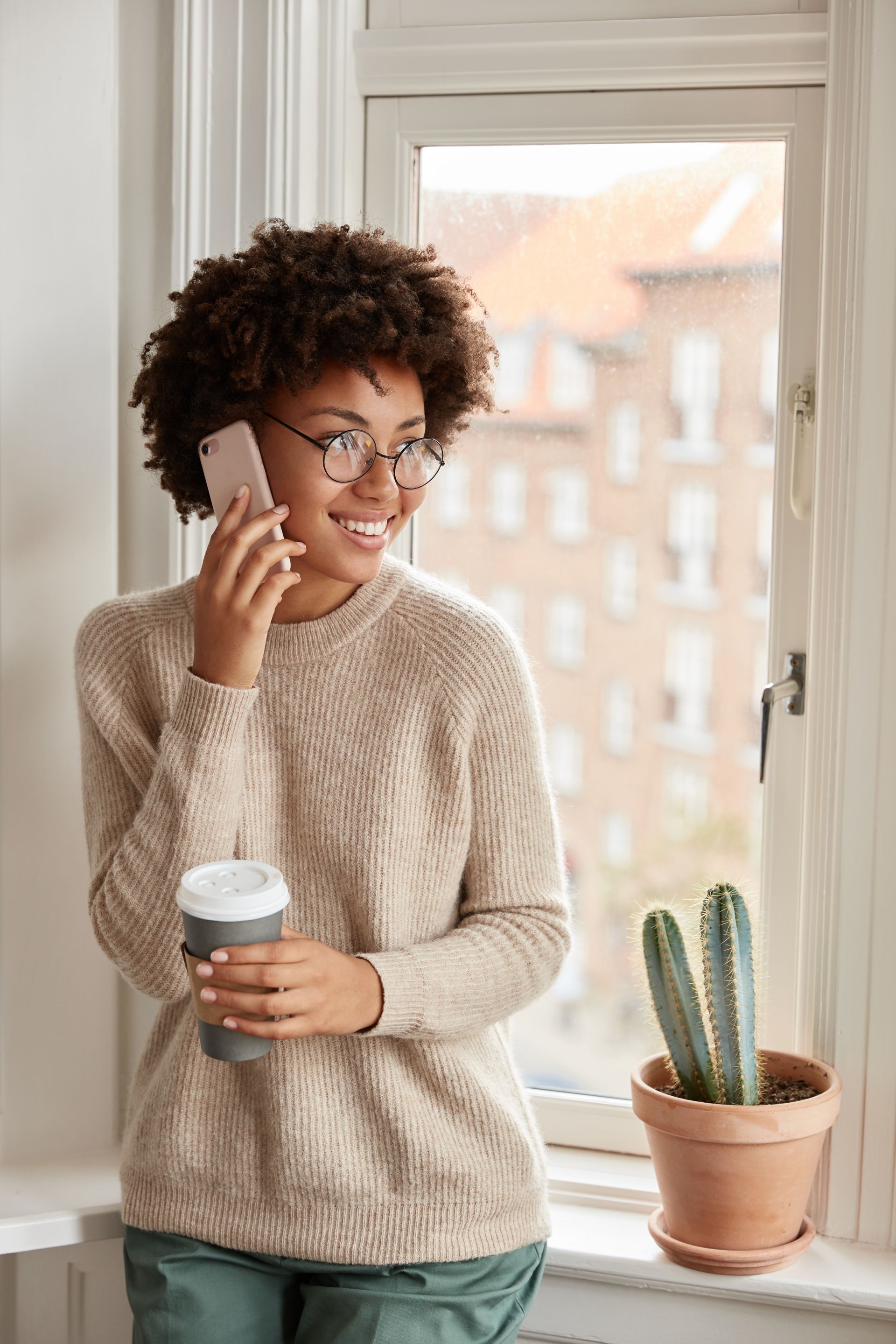 A person with curly hair and glasses holds a coffee cup while smiling and talking on a smartphone by a window.