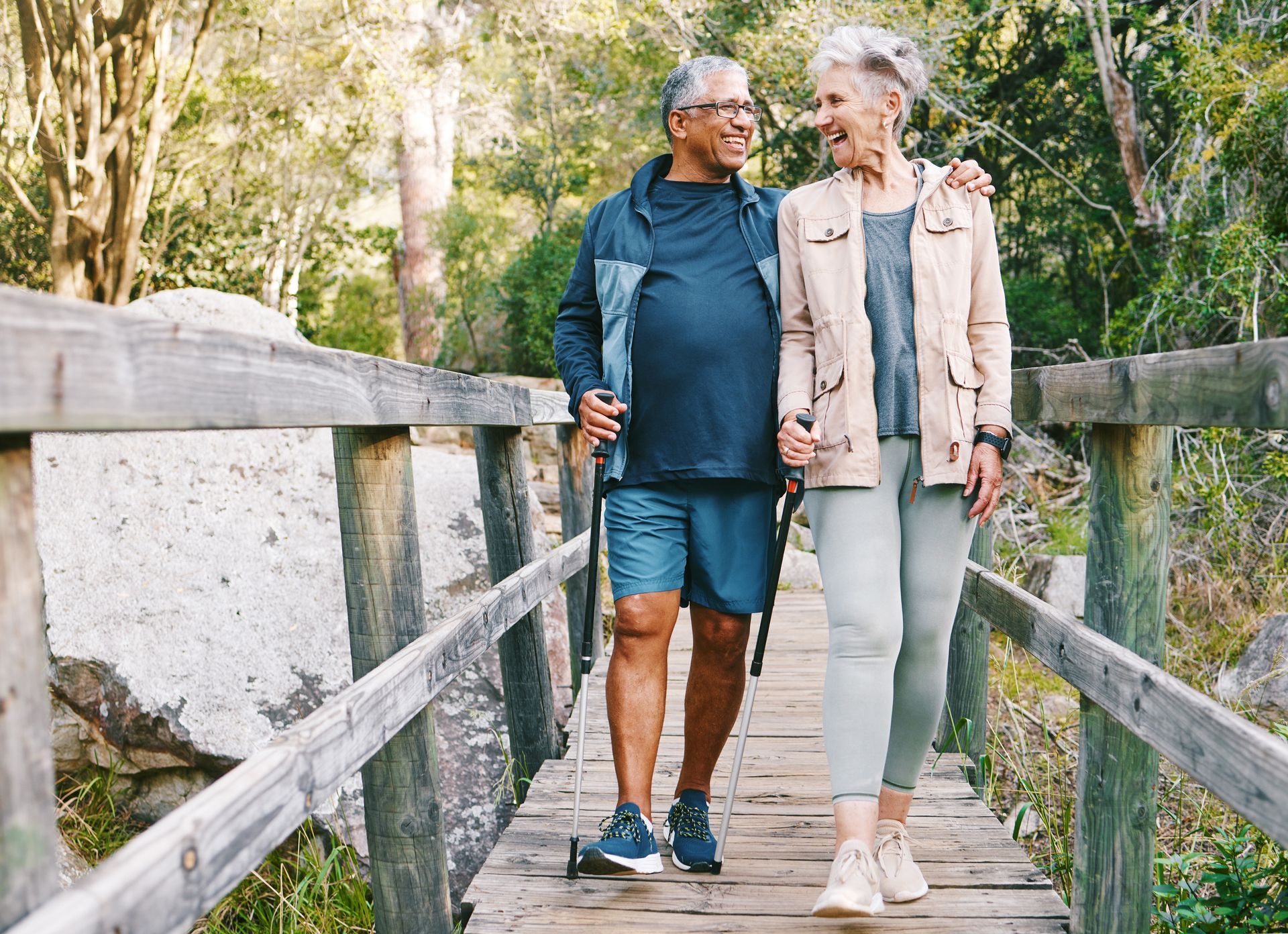 A couple walks across a wooden bridge in a sunlit forest, smiling as they hold walking poles.