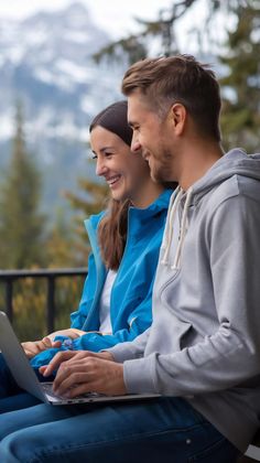 Two people smiling while looking at a laptop on an outdoor balcony with a mountain landscape in the background.