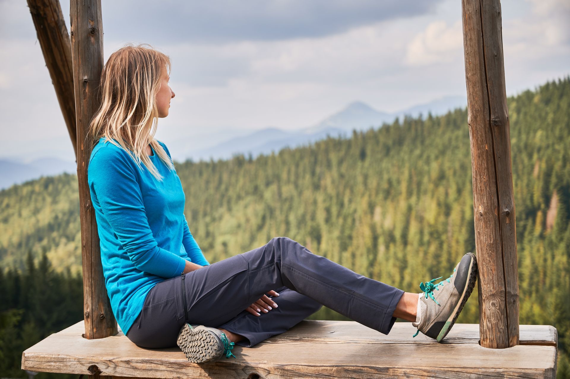 A person with light-colored hair wearing a blue long-sleeve shirt sits on a wooden platform overlooking a mountain forest.