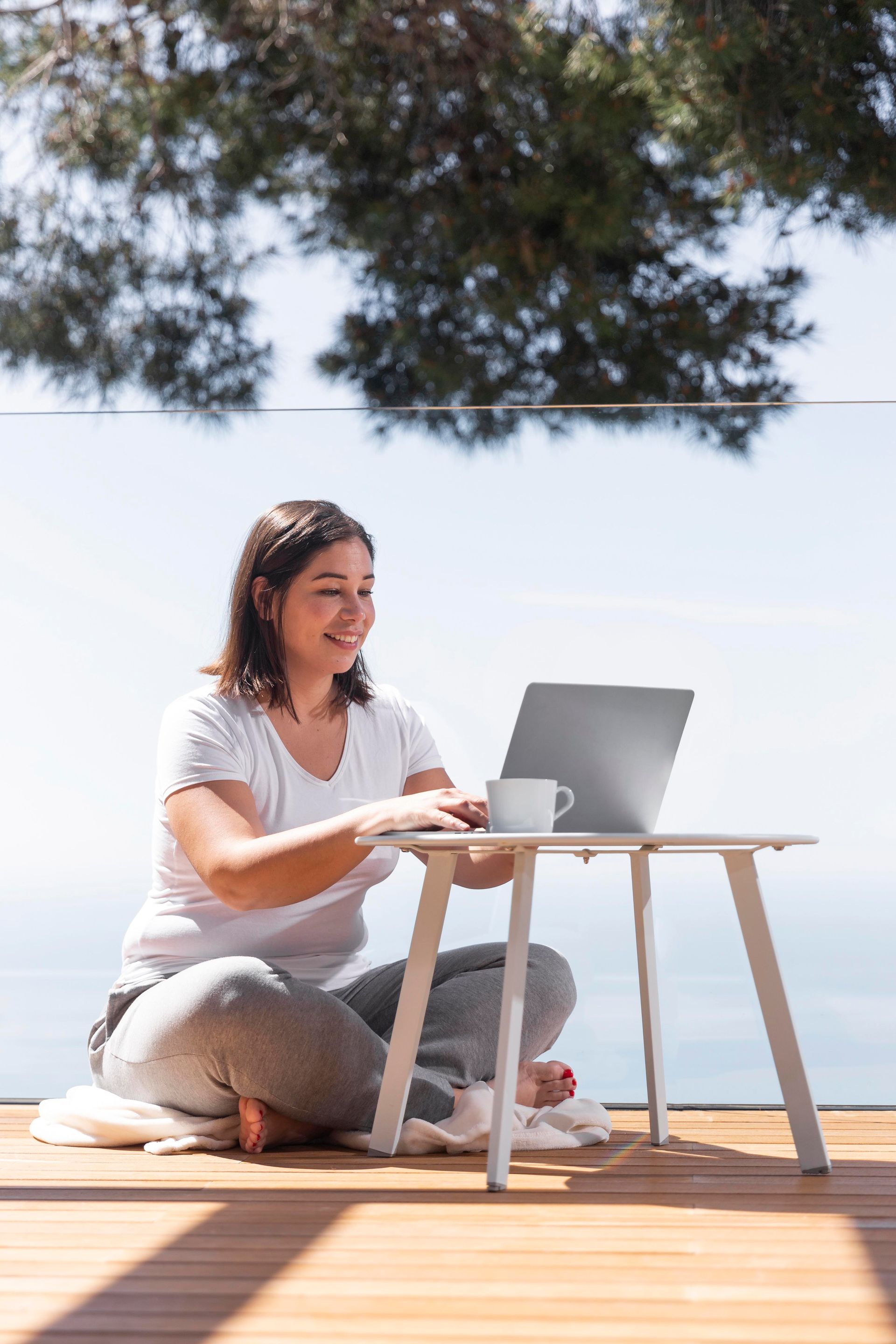 A person sitting cross-legged on a wooden deck, working on a laptop at a small table with a cup nearby, outdoors.