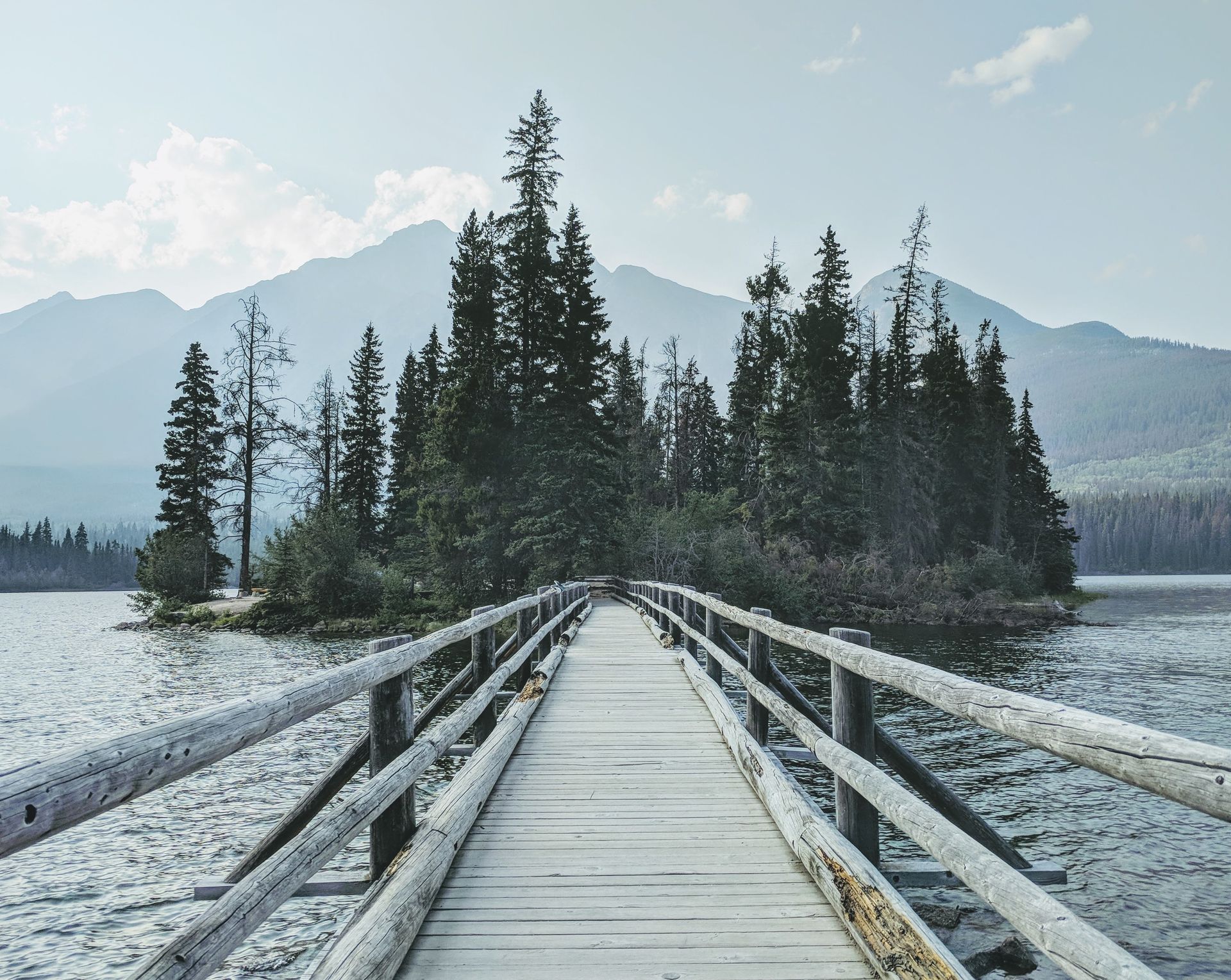 A long wooden footbridge stretches over water toward a forested island set against misty mountain peaks.
