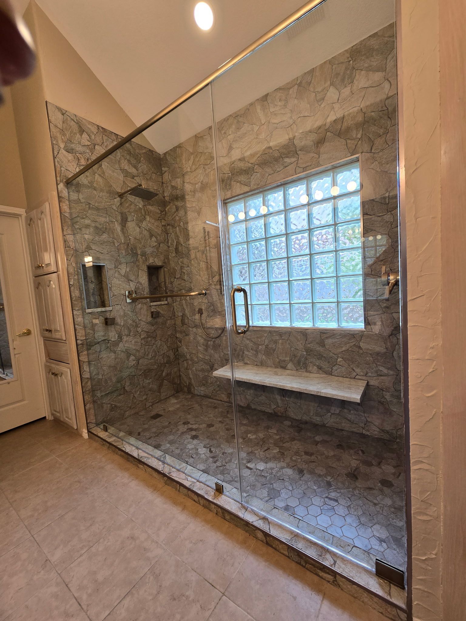 A modern shower with gray tiled walls and a glass door. A window of glass blocks is visible.