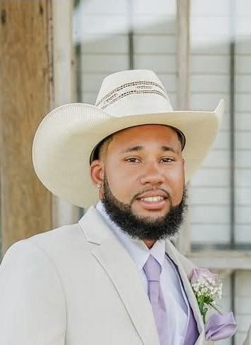 Man wearing a cowboy hat, suit, and tie, smiling. He has a beard and is standing outside a building.