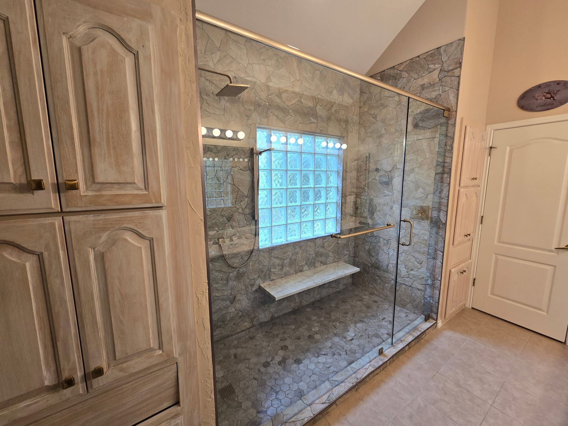 A bathroom with a glass shower, built-in bench, and a glass block window. Light wood cabinetry and a closed door are visible.