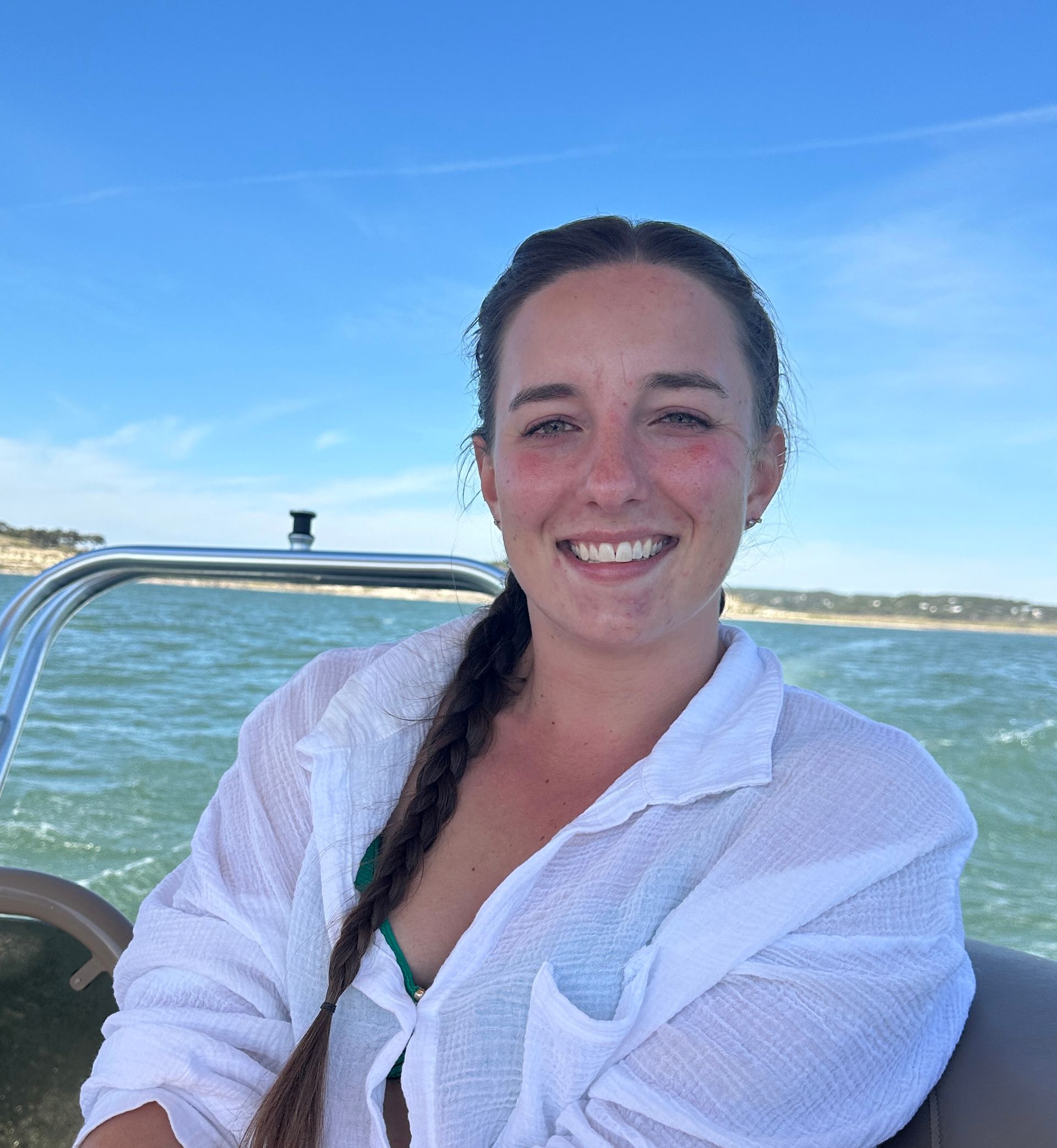 Woman on a boat, smiling at the camera. She has long hair in a braid, wearing a white shirt, with a blue sky and water in the background.